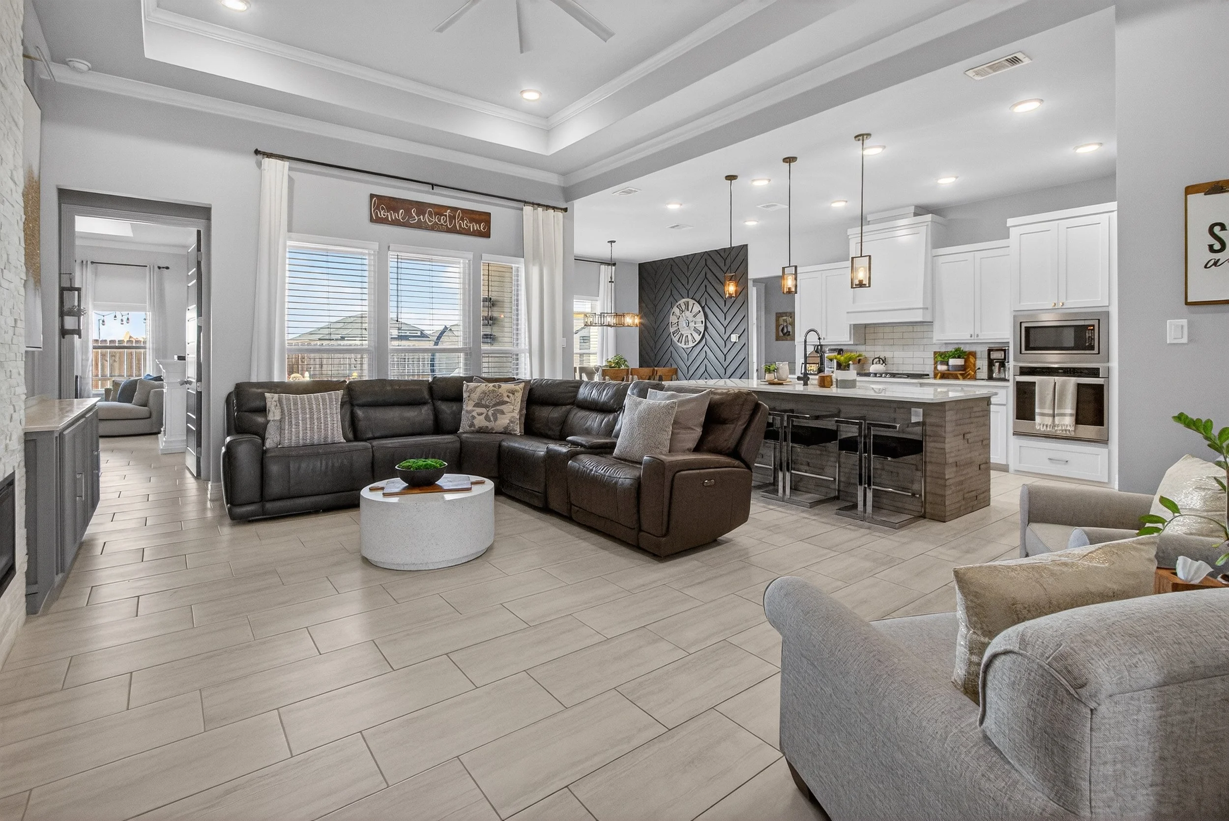 Open concept living room and kitchen with white cabinets, modern appliances, a dark accent wall with a large clock, a black leather sofa, and a dining table with chairs.