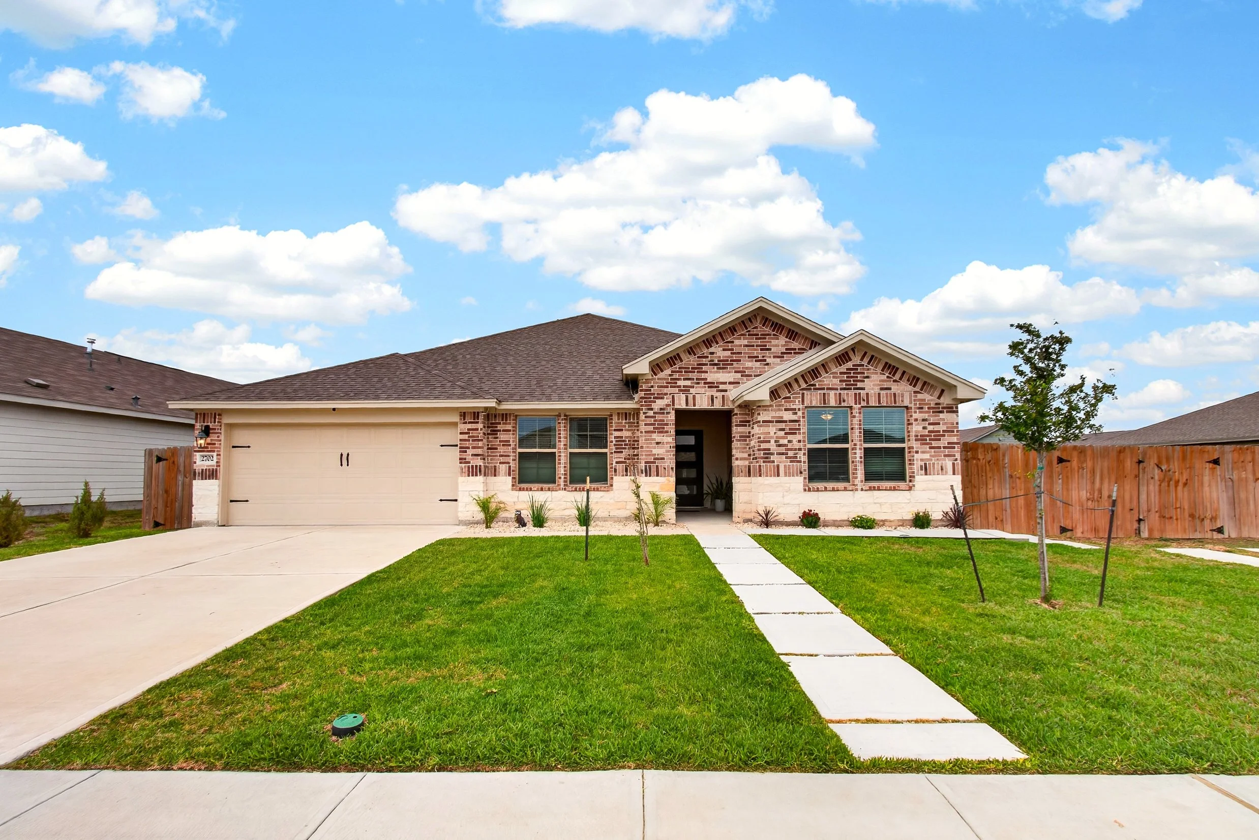 A new house with a brick exterior, a driveway, a small front yard with grass and young trees, and a blue sky with scattered clouds.