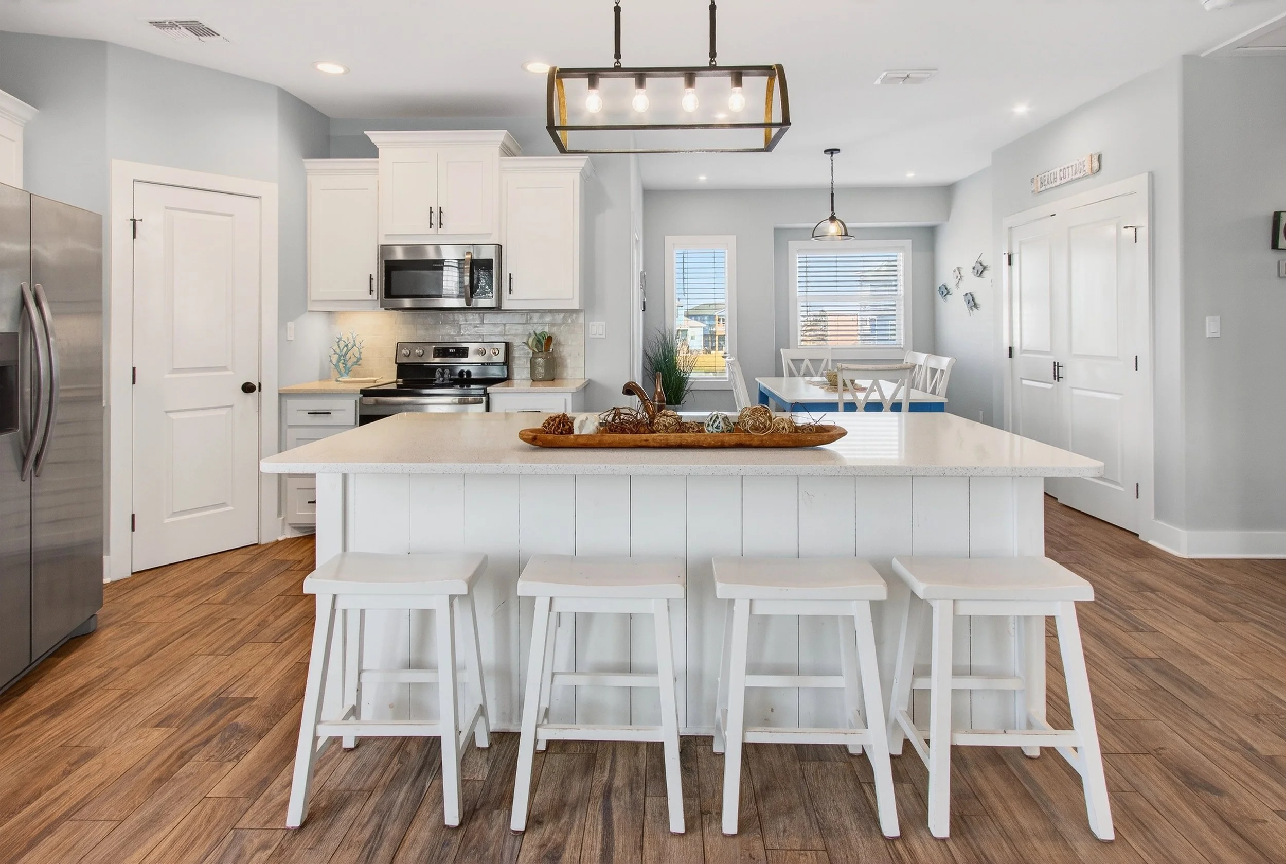 A modern kitchen with a white island, five white stools, a light wood floor, white cabinetry, and a dining area with a white table and chairs. There are windows with blinds and a light fixture. Decorations include a wooden tray with balls of twine on