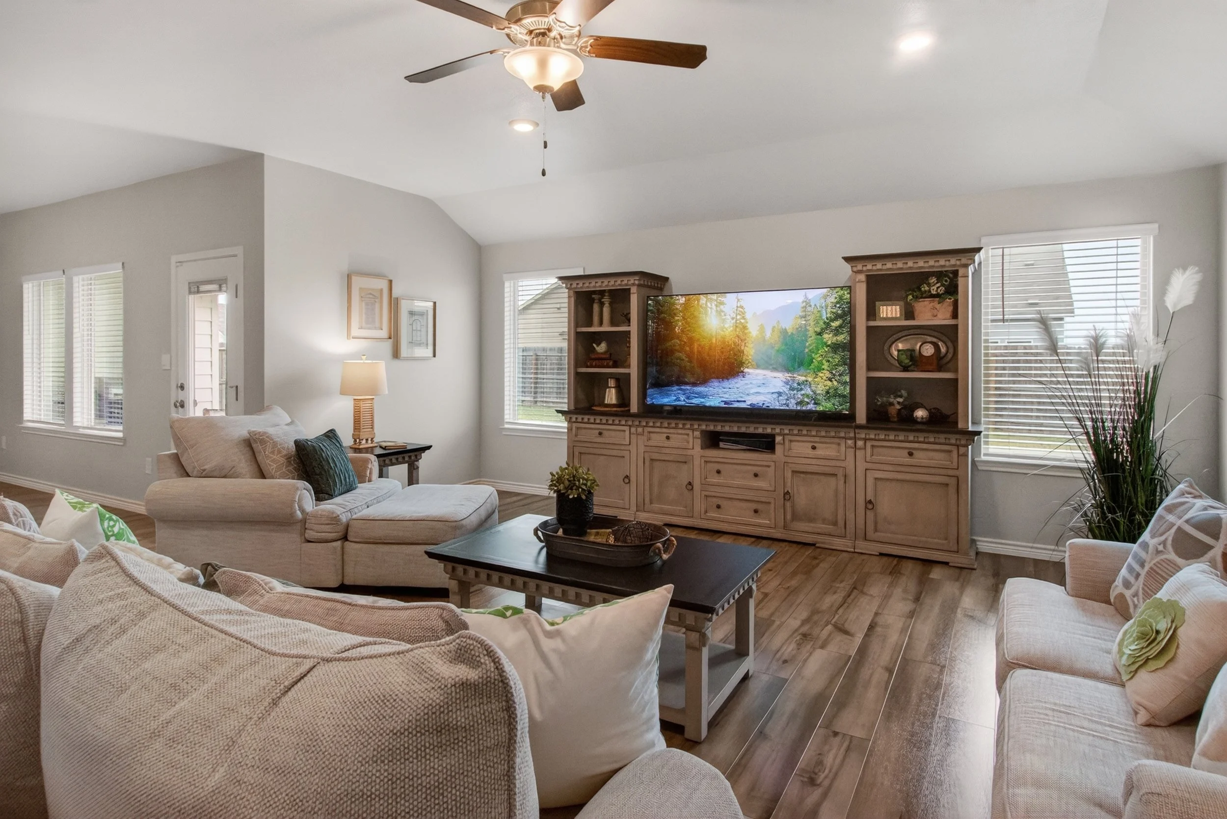 Living room with beige sofas, a black coffee table, a wooden entertainment center with a television displaying a nature scene, a ceiling fan, large windows with blinds, and various decorative items.