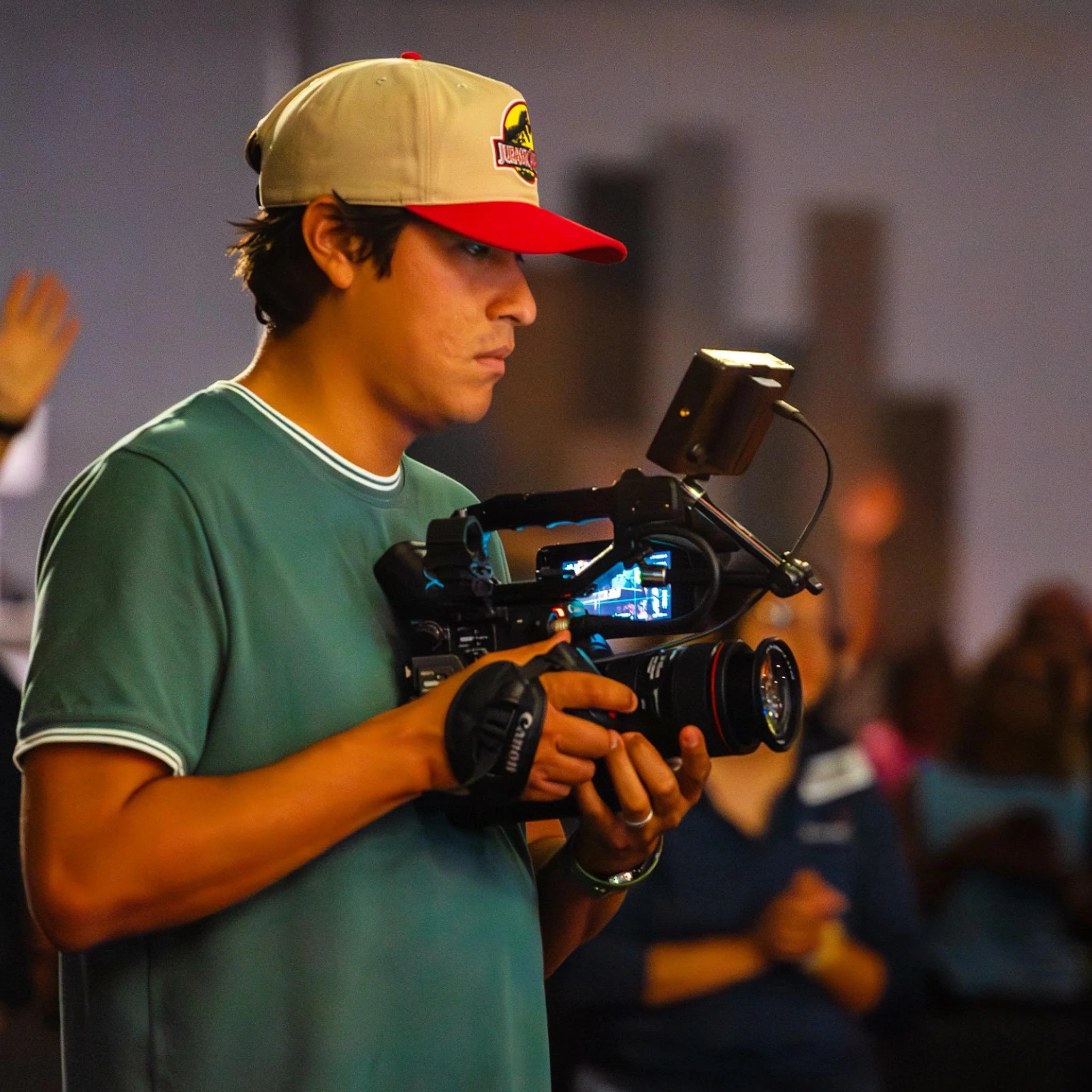 Man wearing a Jurassic Park cap holding a professional video camera during an event.