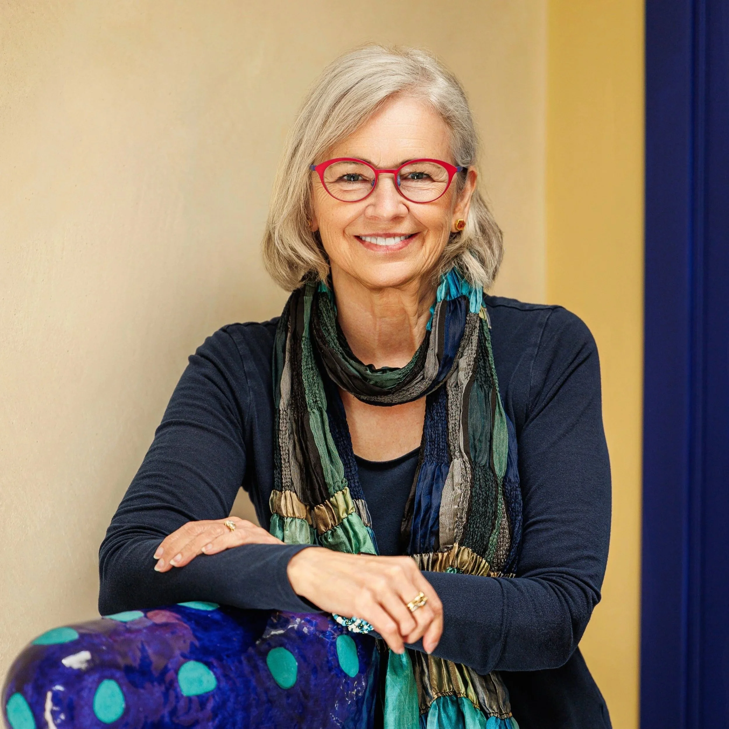 A smiling elderly woman with gray hair wearing red glasses, a multicolored scarf, and a dark blue top, standing against a yellow and blue wall.