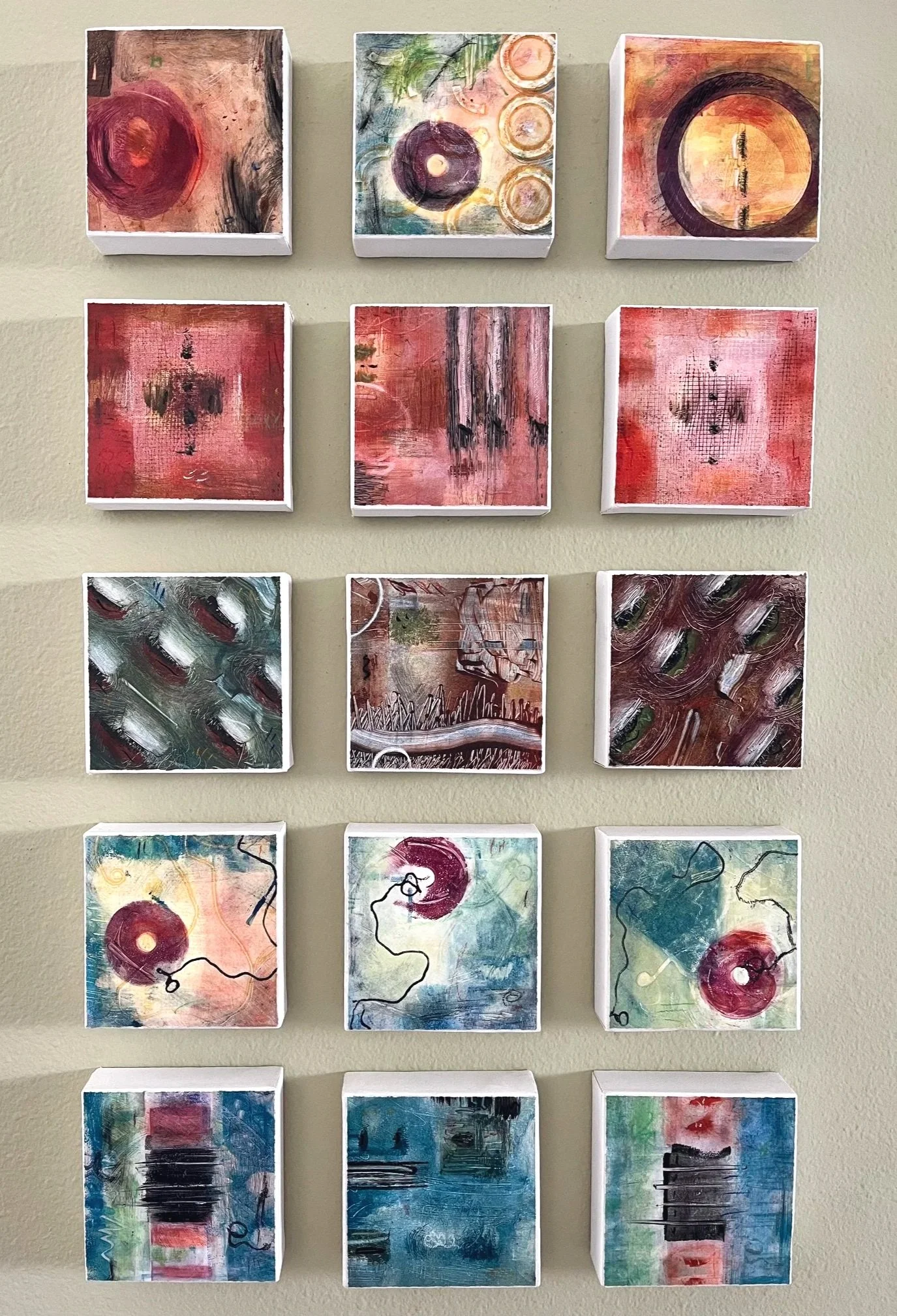A wall display of nine abstract mixed media paintings arranged in a grid with three rows and three columns. Each piece features vibrant, textured, and layered designs with various shapes and brushstrokes.