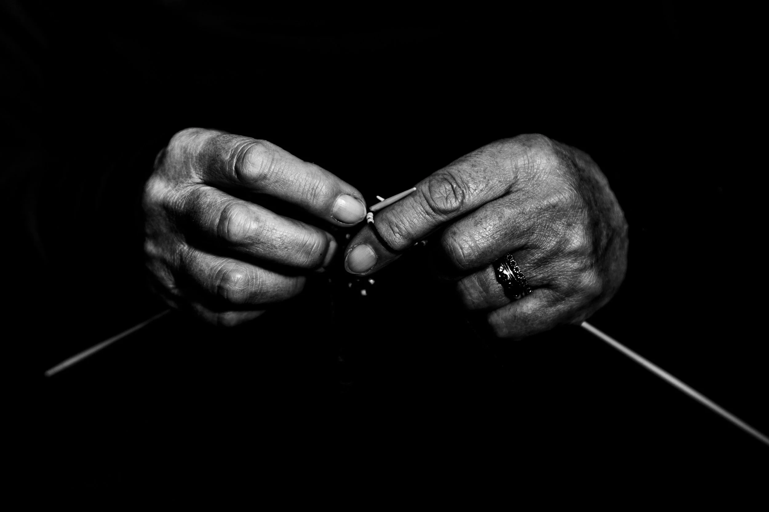 Black and white close-up of hands holding a needle and thread, with a focus on detailed skin and fingers, dark background.