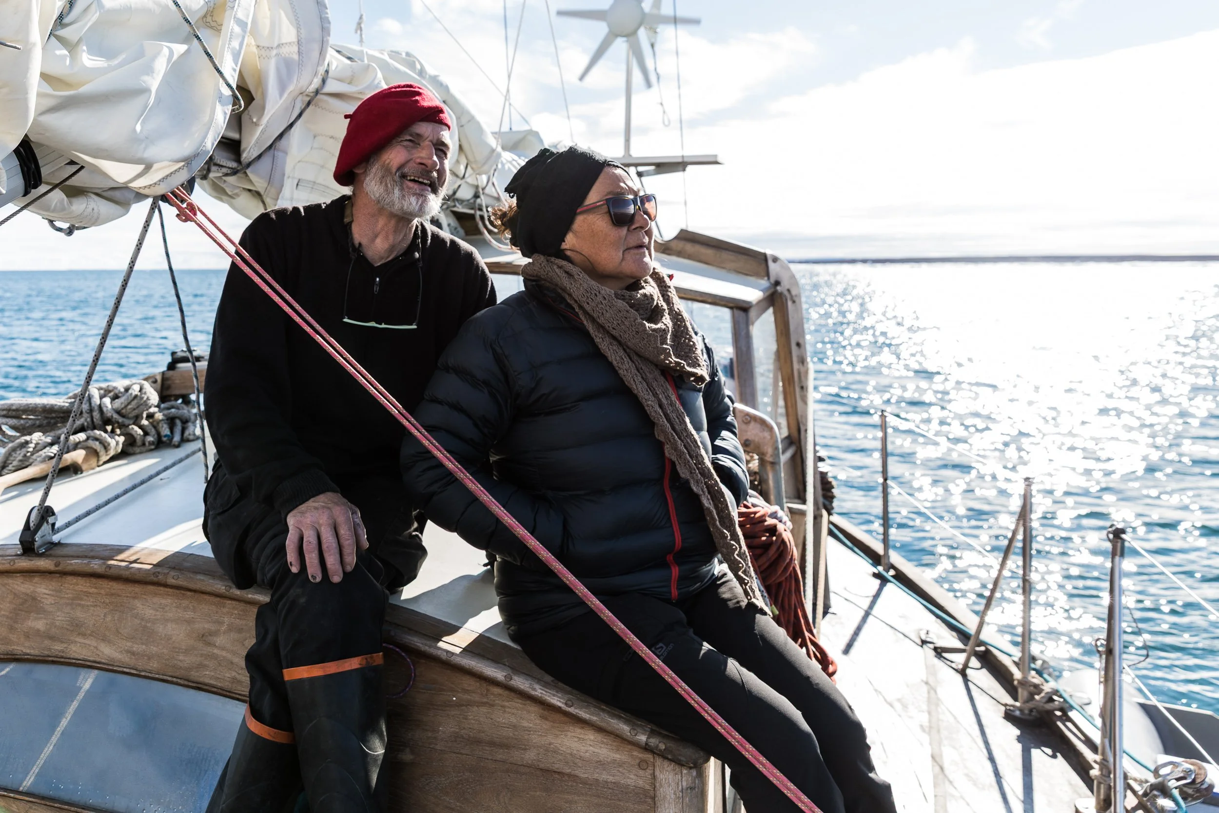 Two people sit on the side of a sailboat, enjoying the view of the ocean with the sun shining on the water, amid a partly cloudy sky.
