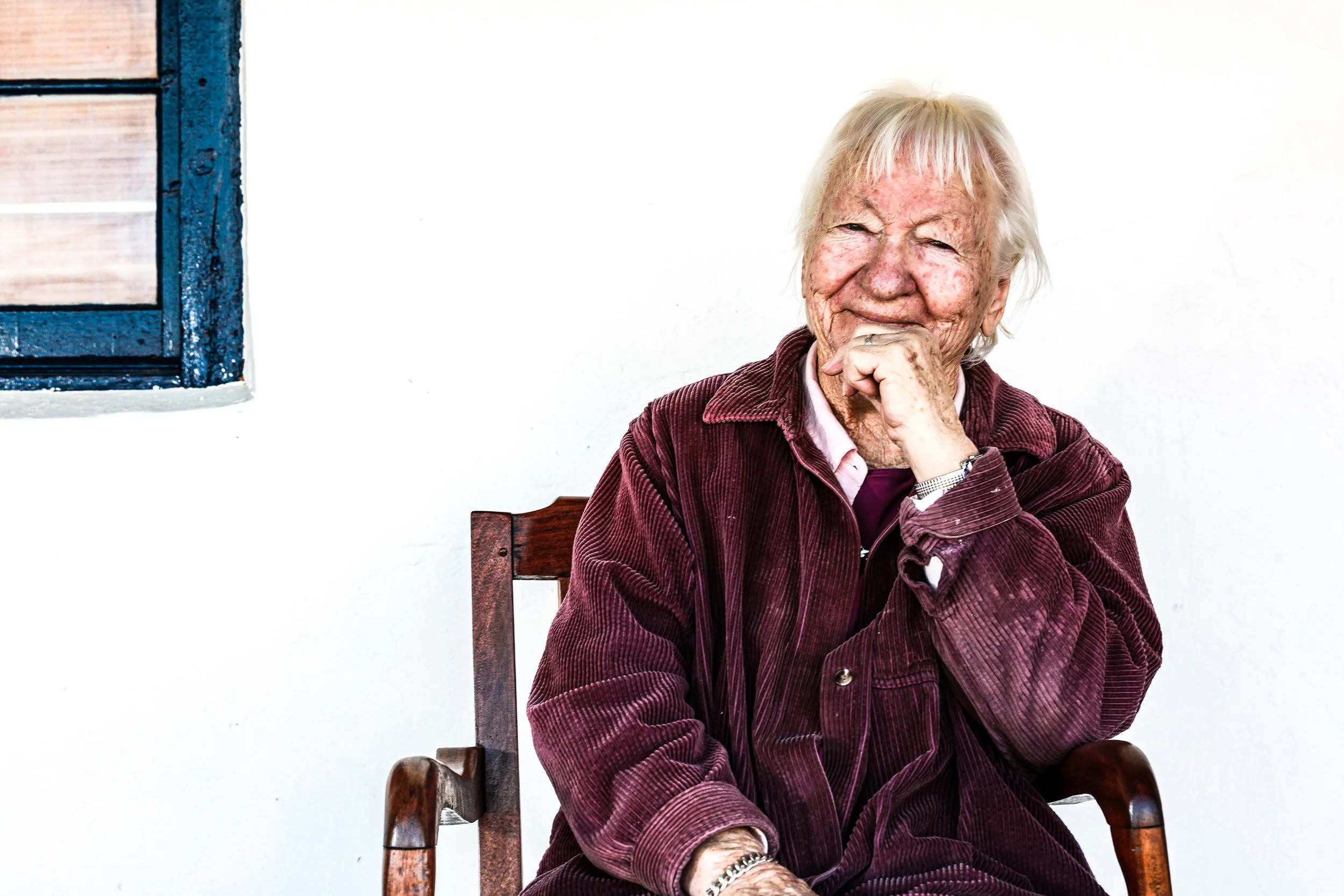 An elderly woman with white hair and wrinkles, sitting on a wooden chair, smiling and resting her chin on her hand, wearing a maroon corduroy jacket and a pink shirt, in front of a white wall with a small blue window on the side.
