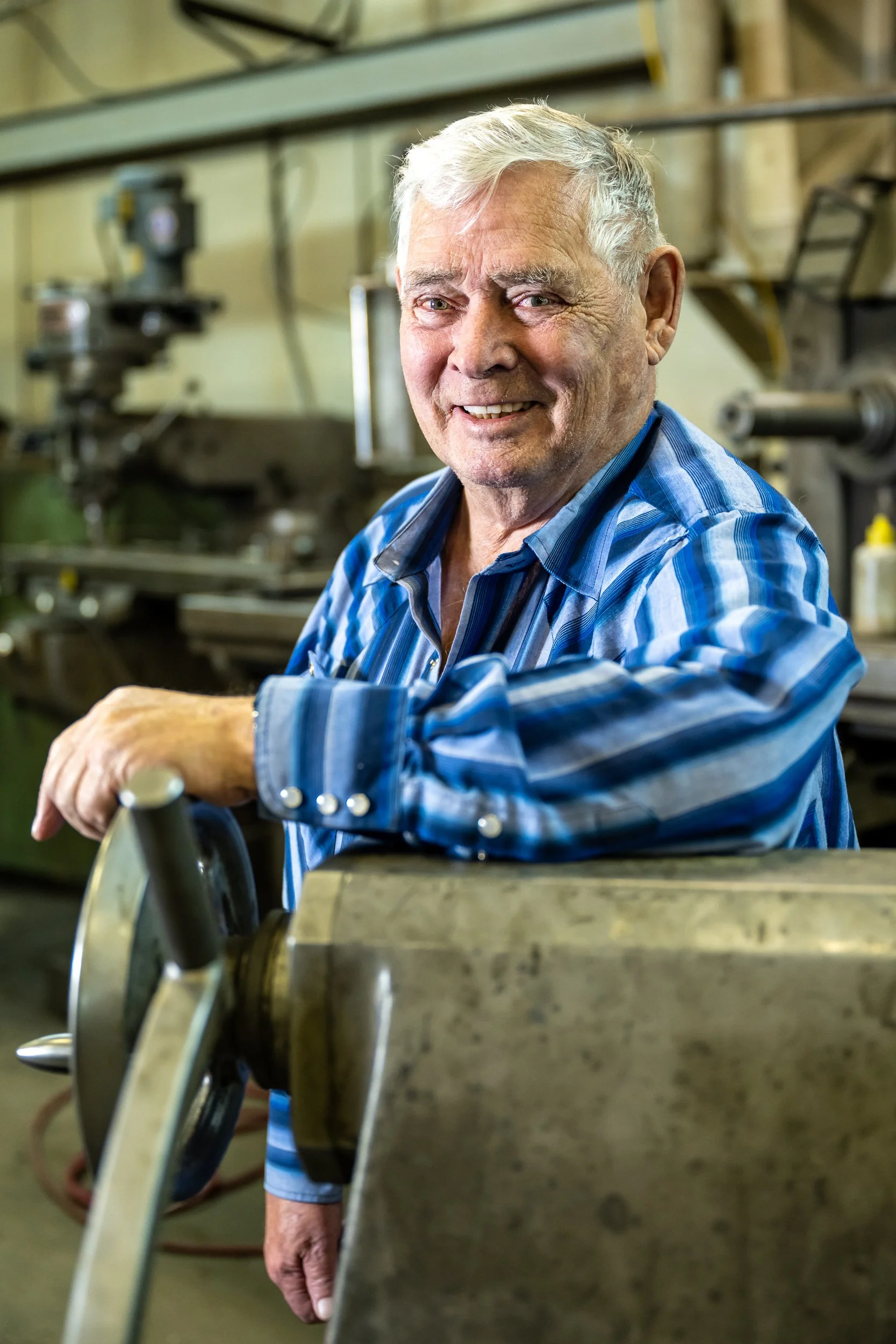 An elderly man with gray hair smiling, working in a workshop with machinery and tools around him, wearing a blue plaid shirt.