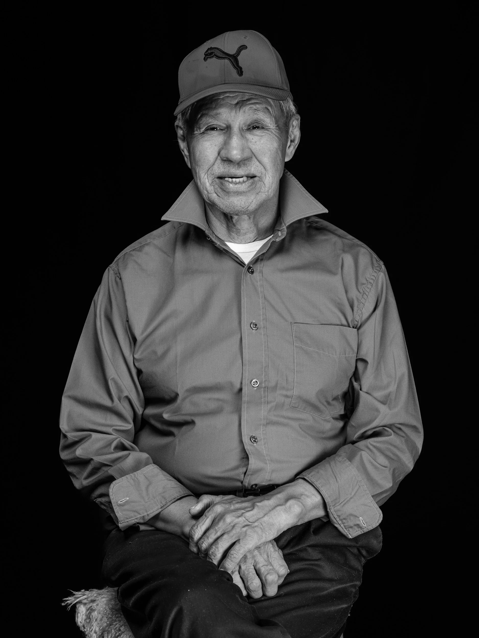Black and white photo of an elderly man wearing a baseball cap with Puma logo and a button-up shirt, sitting against a black background, with hands clasped over his lap.