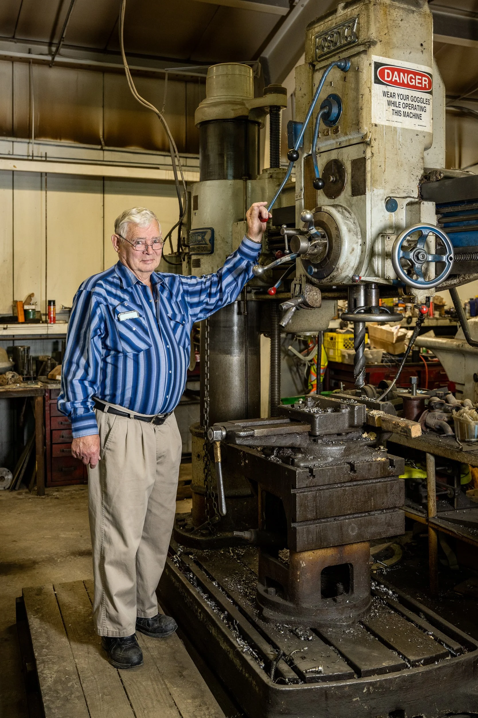An older man in glasses and a blue striped shirt stands in a workshop, operating a large industrial machine, with signs including a red danger sign on it.