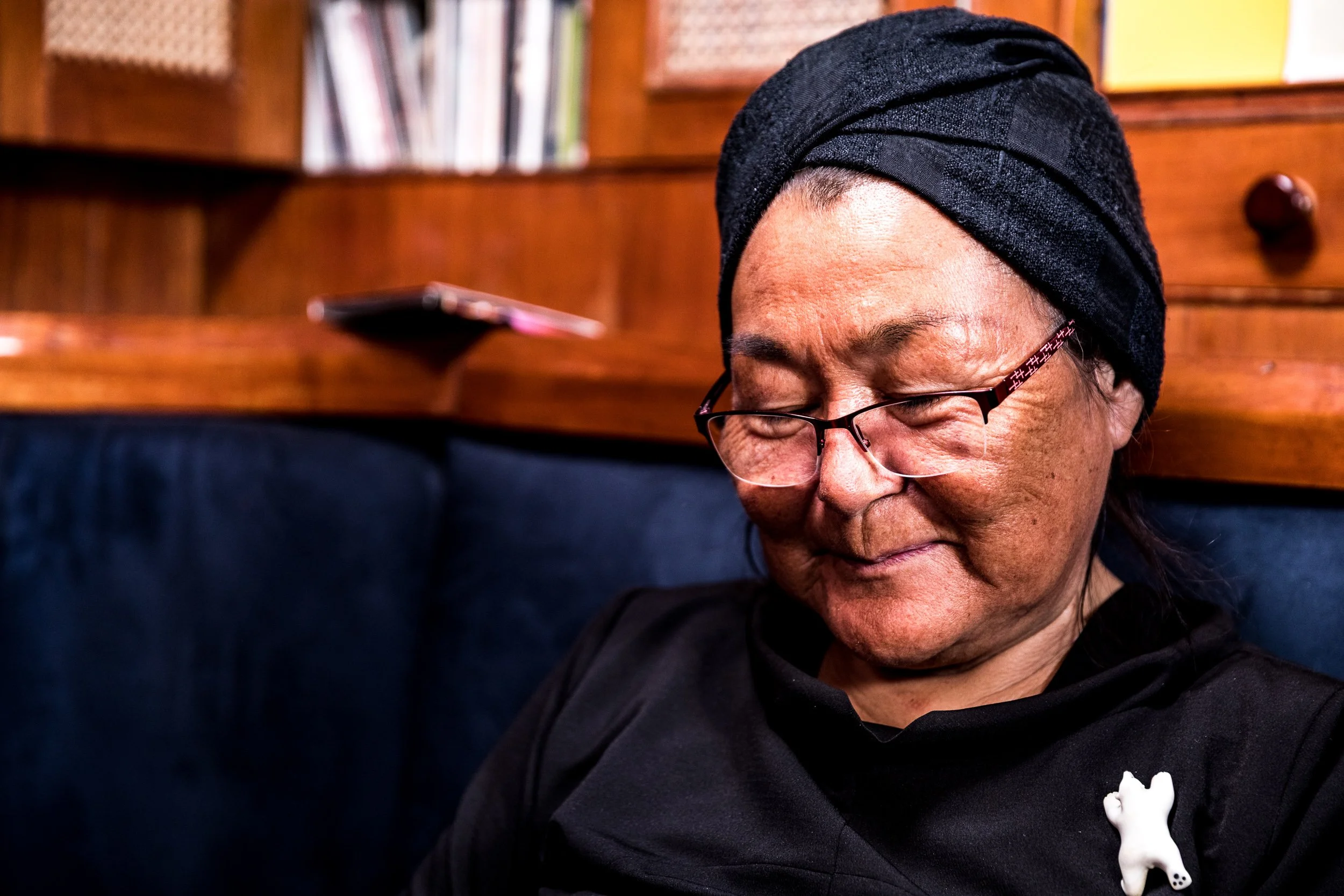 A woman with glasses, dark head wrap, and black shirt smiling and looking down, sitting in front of a wooden bookshelf.