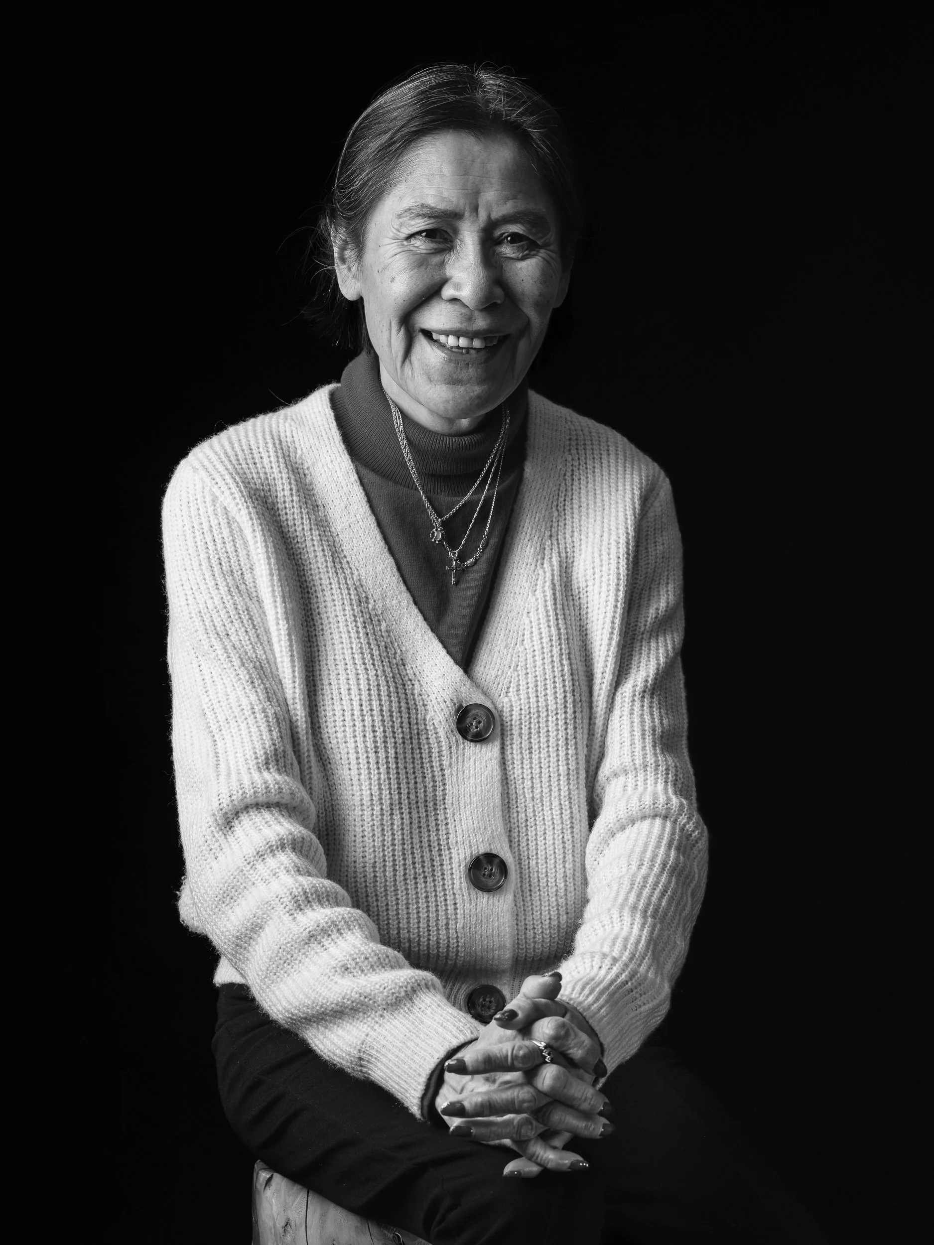 Black-and-white portrait of an elderly woman smiling, wearing a knitted cardigan and layered necklaces, with hands gently clasped, against a dark background.