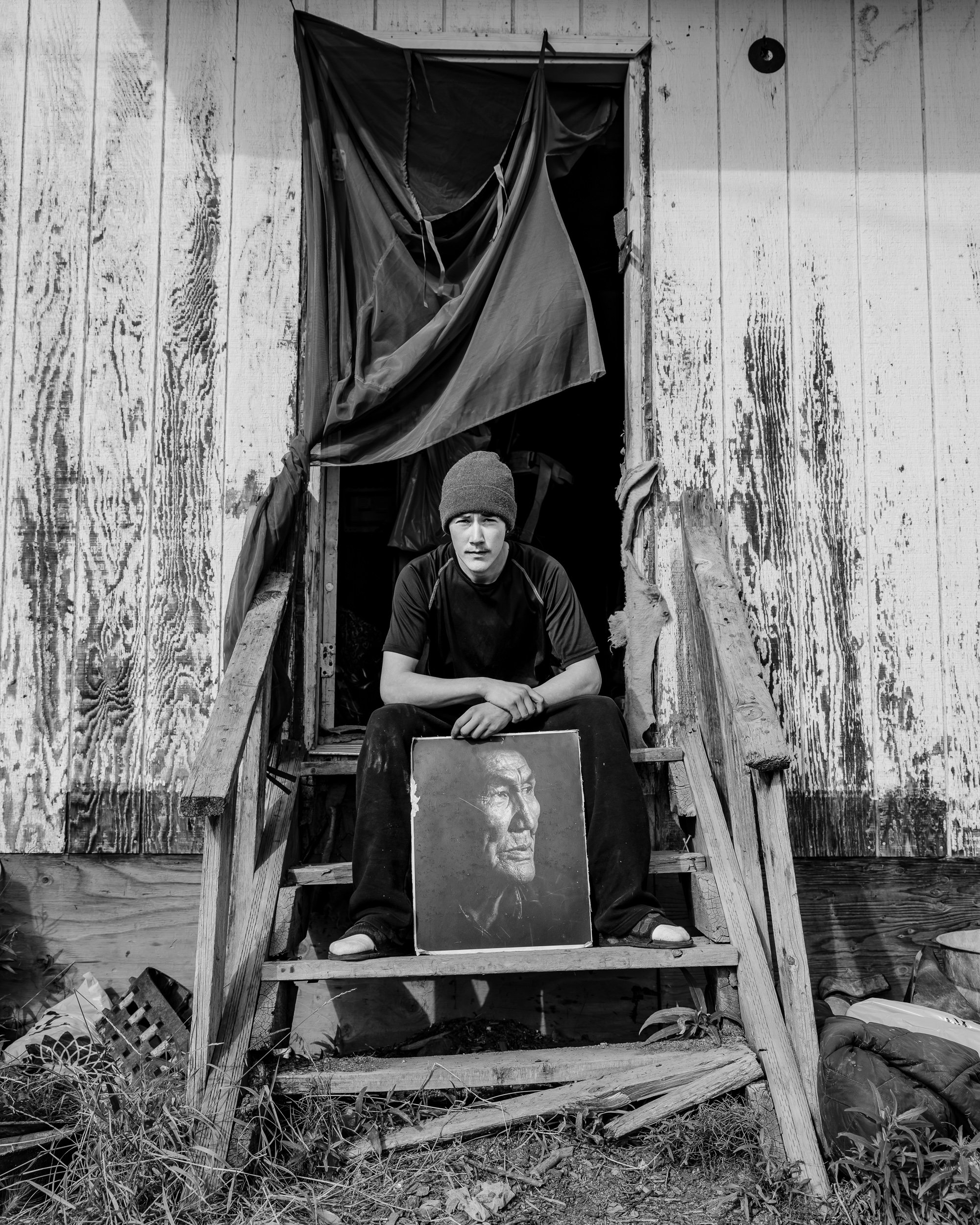 A young man wearing a beanie sits on the steps of a dilapidated wooden house, holding a portrait of an elderly woman with a serious expression. The house appears abandoned and in disrepair, with torn fabric hanging over the doorway and scattered debr