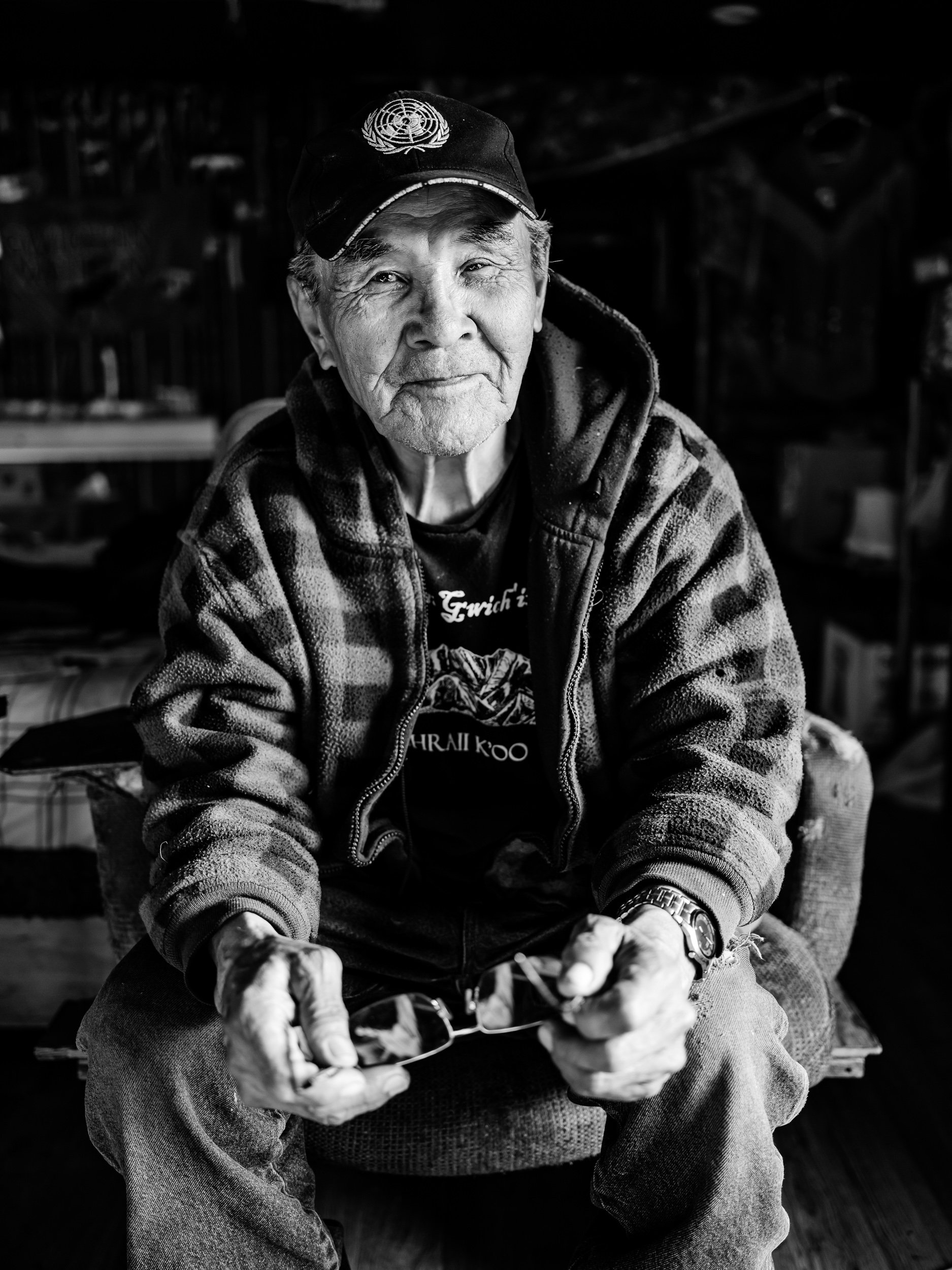 An elderly man wearing a cap with the UN logo, a hoodie, and a watch, sitting indoors, holding a bowl with food, looking at the camera.