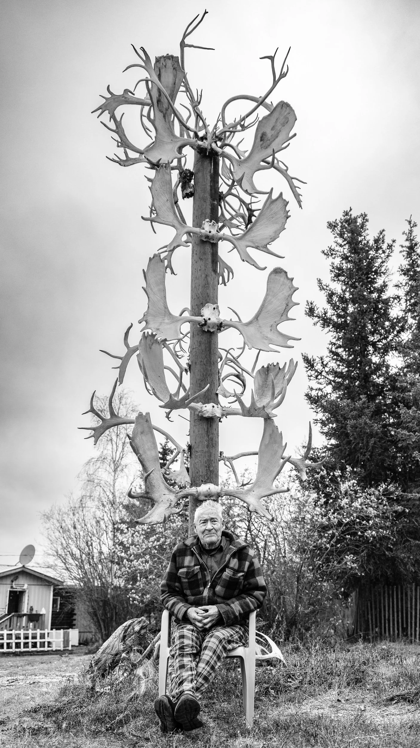 An elderly man sitting in a plastic chair outdoors, with a large antlered animal skull mounted on a pole behind him. The scene appears to be in a backyard with trees and a house in the background.