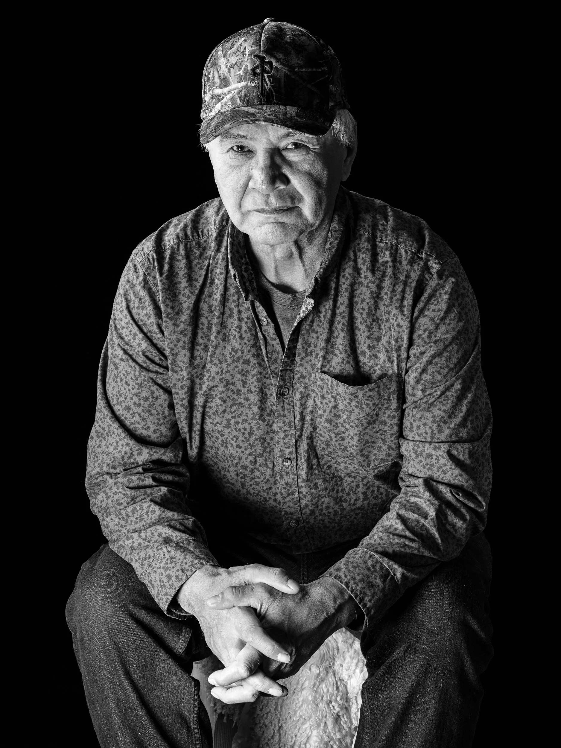 Black and white portrait of an older man wearing a camo baseball cap and a patterned shirt, sitting with his hands clasped together.
