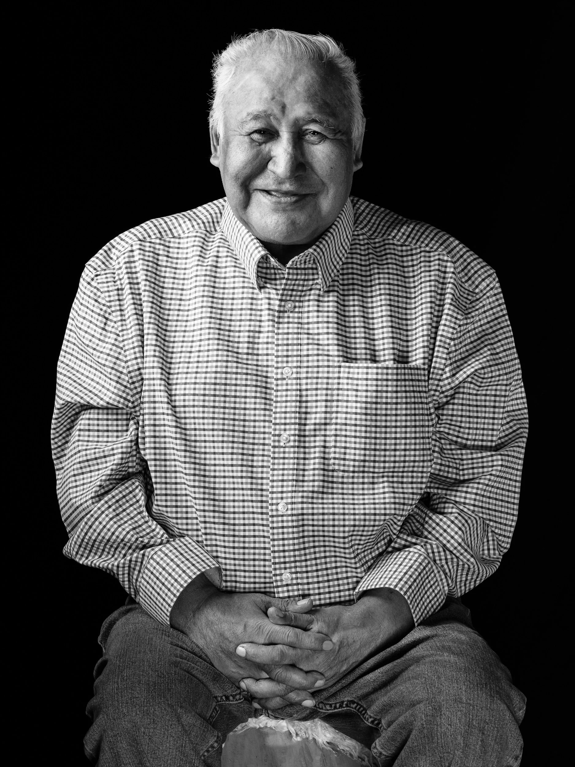 An elderly man with gray hair, wearing a checked shirt, with hands clasped on his lap, looking at the camera, sitting in front of a plain black background.