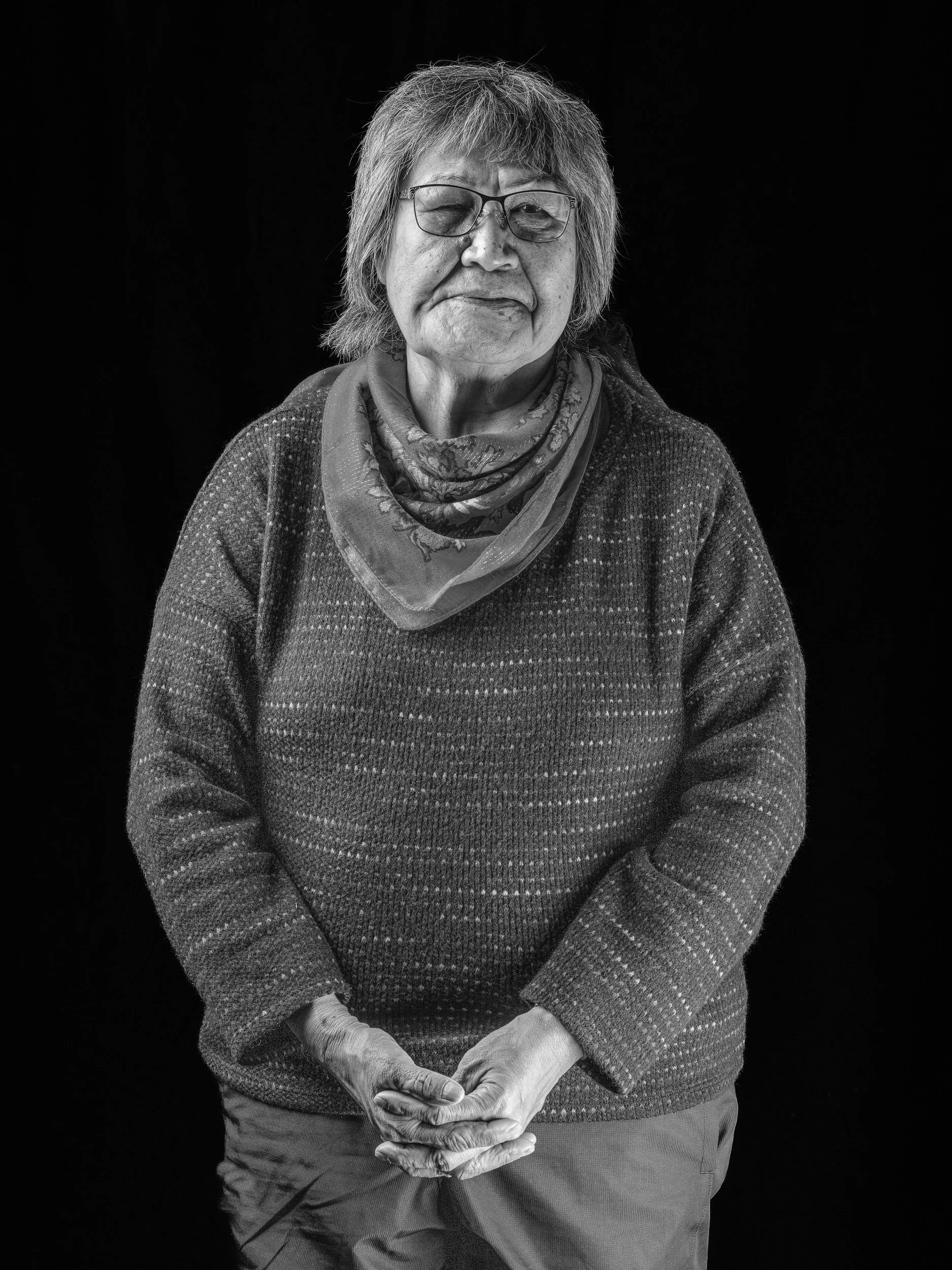 A black and white portrait of an elderly woman with glasses, wearing a patterned scarf and a knitted sweater, against a dark background.