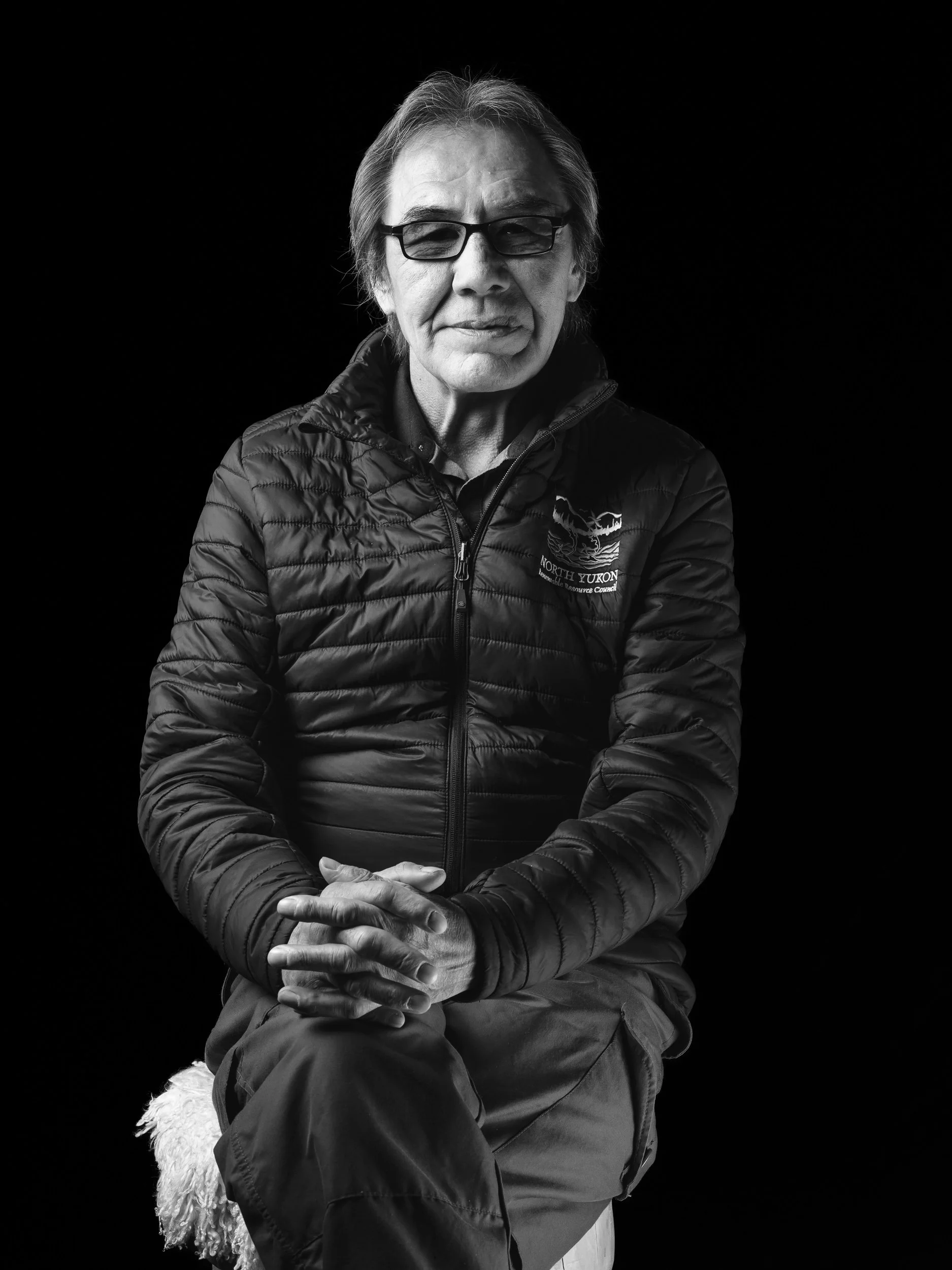 Black and white portrait of an elderly woman with glasses, wearing a North Yukon jacket, sitting with her hands clasped on her lap, against a dark background.