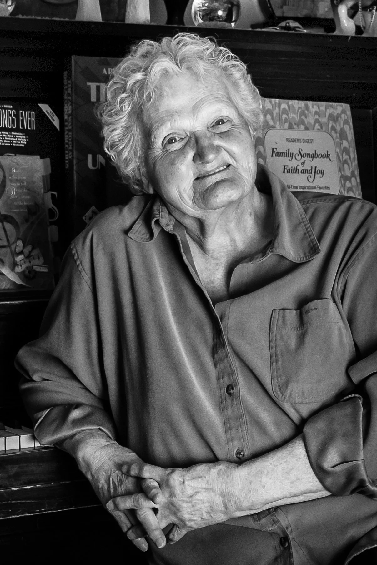Older woman with curly hair sitting in front of a bookshelf, wearing a button-up shirt, with her hands folded in her lap, looking at the camera with a slight smile.