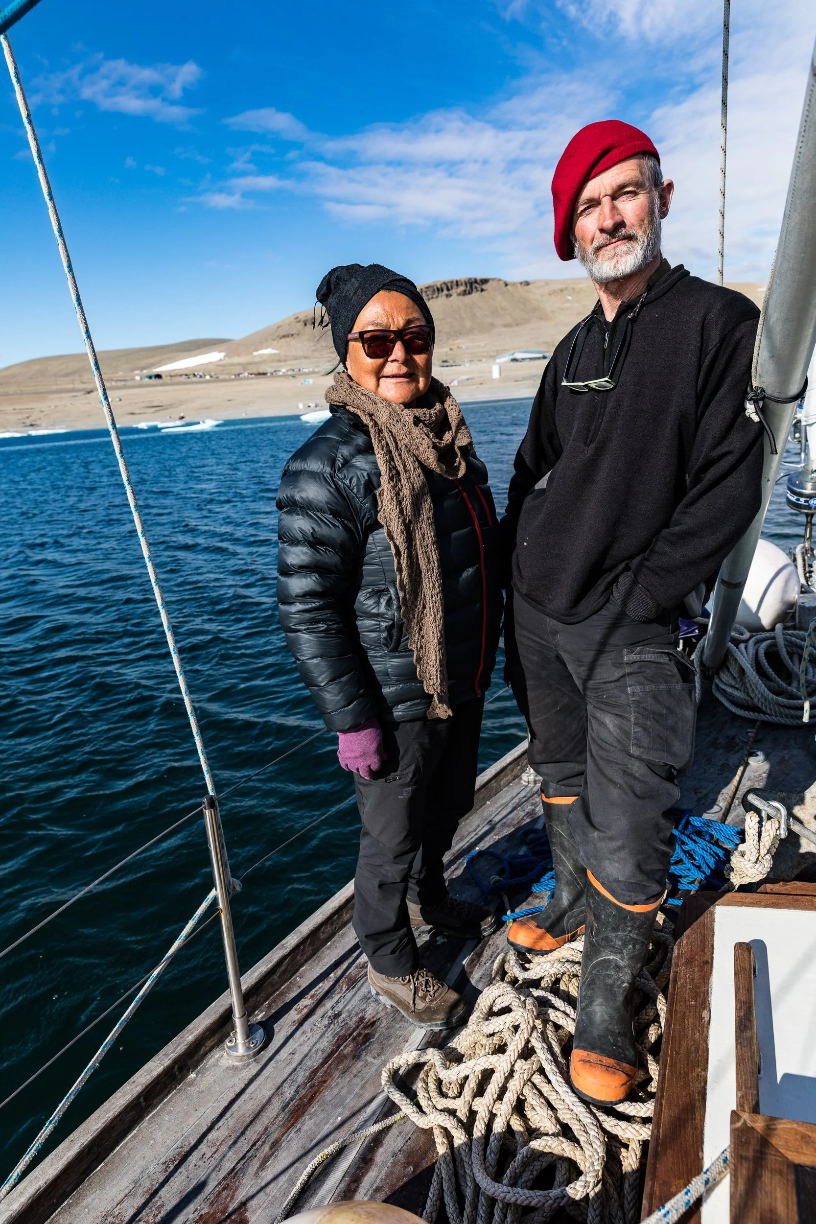 Two people standing on a boat with water and land in the background, dressed for cold weather, one wearing a red beret and the other wearing a black beanie, with some ropes on the deck.