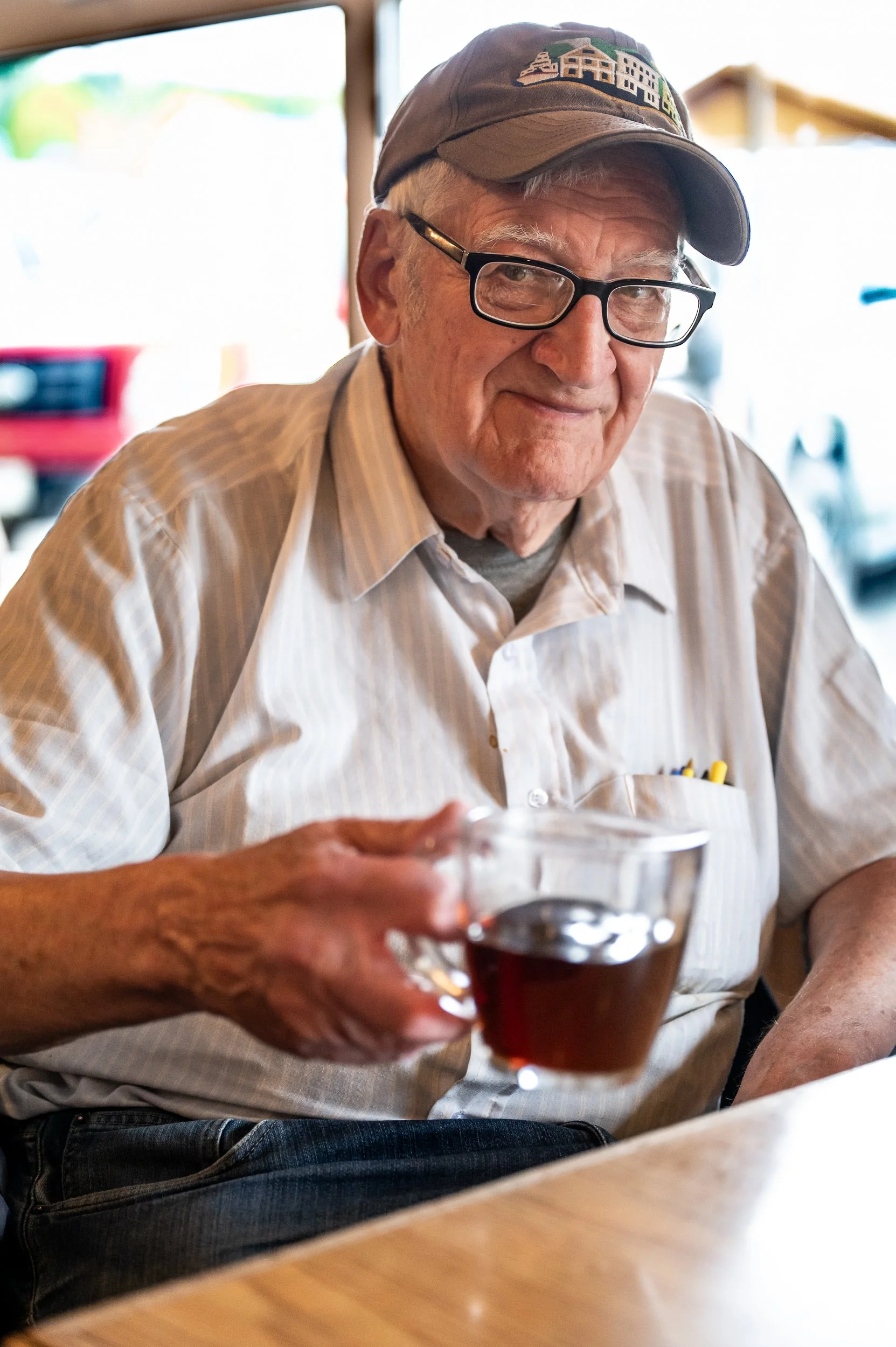 An elderly man wearing glasses and a baseball cap, smiling softly, holds a glass of red wine in a cozy indoor setting.