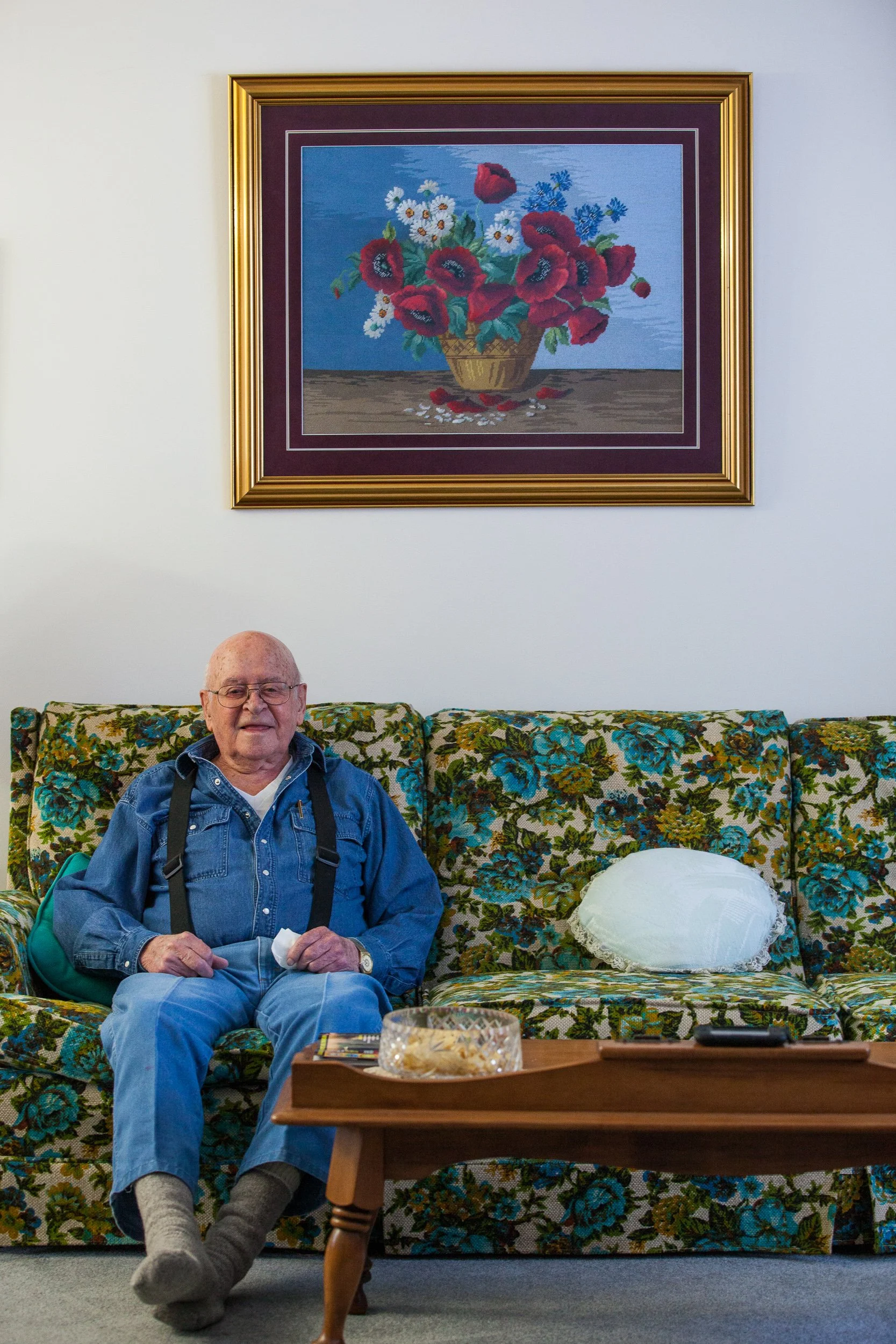 An elderly man with glasses, wearing a blue denim shirt and gray socks, is sitting on a floral-patterned couch with a gold and green design. He is holding a tissue in his right hand and a tissue box is on the table in front of him. Behind him on the 
