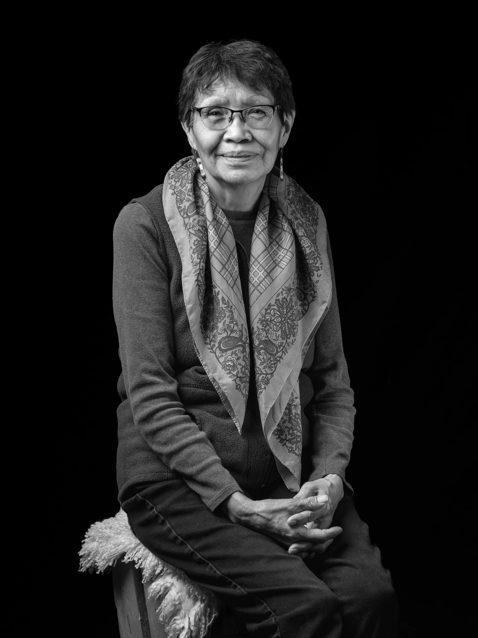 A black and white portrait of an older woman with short hair, glasses, and hoop earrings, sitting on a chair with a fluffy cushion, wearing a paisley scarf and a zip-up jacket against a dark background.