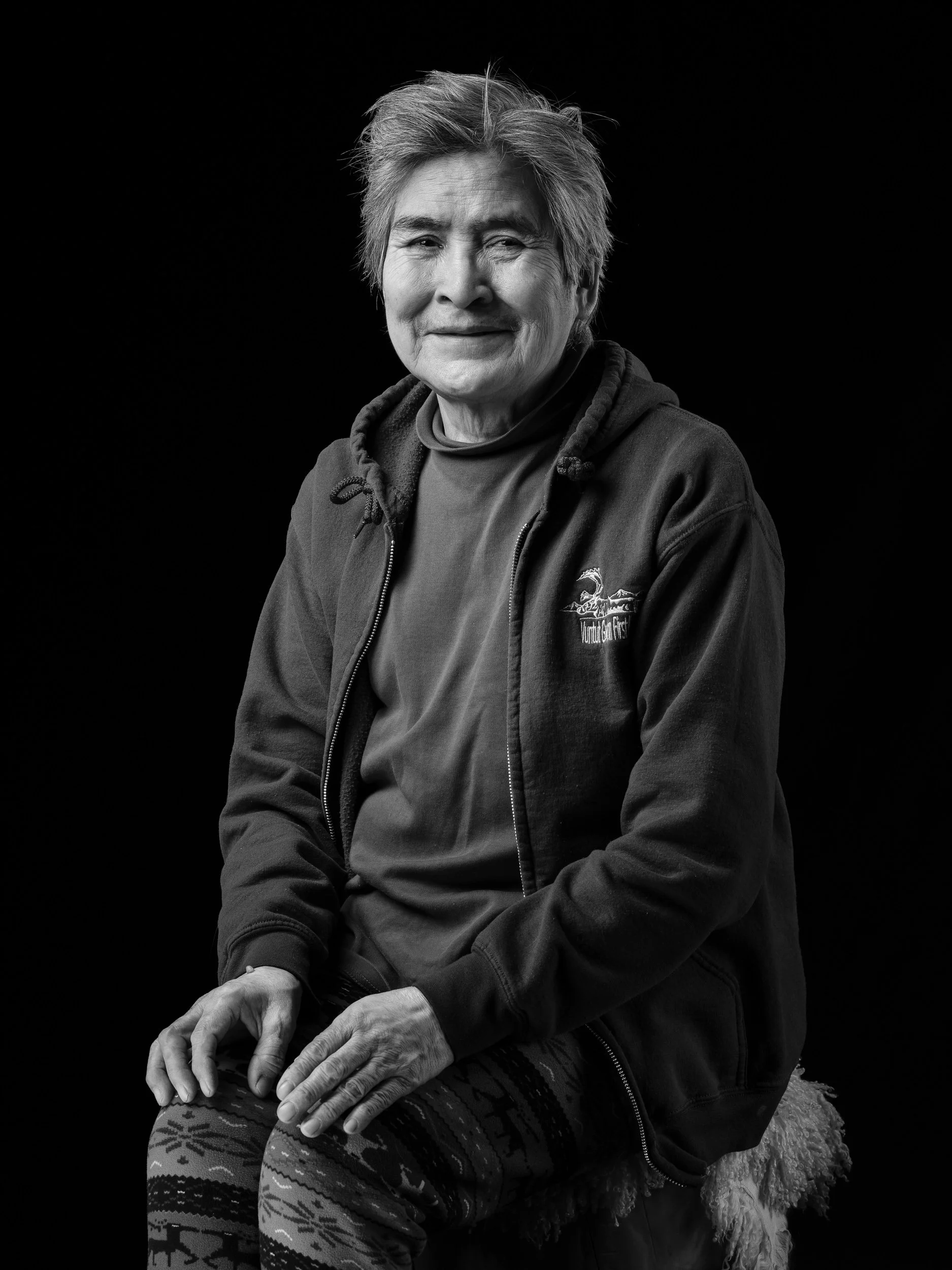 A black and white portrait of an elderly woman with short gray hair, smiling gently, wearing a zip-up hoodie with a logo, and patterned leggings, sitting against a dark background.