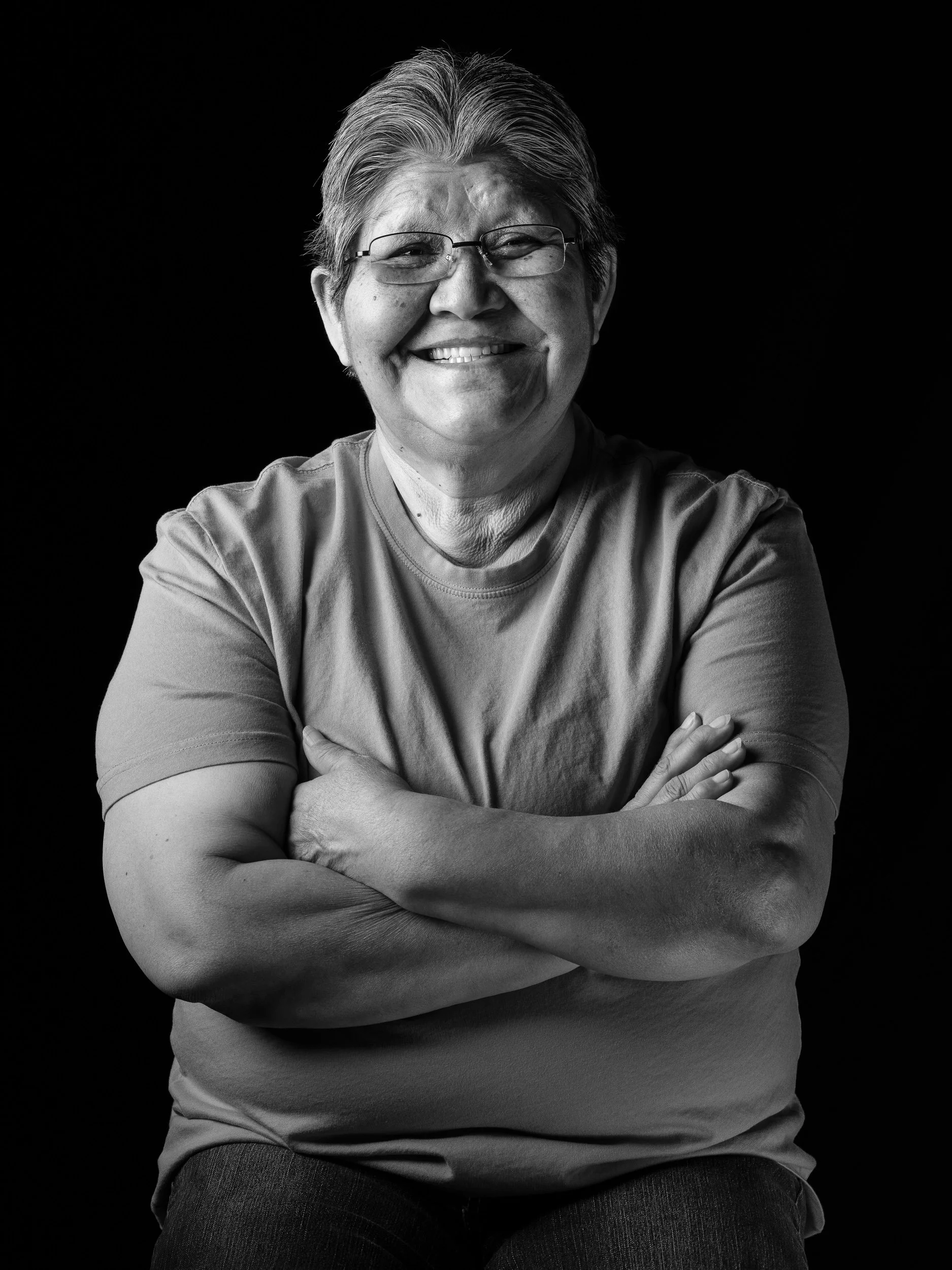 Black and white portrait of a smiling middle-aged woman with glasses, crossing her arms, against a dark background.