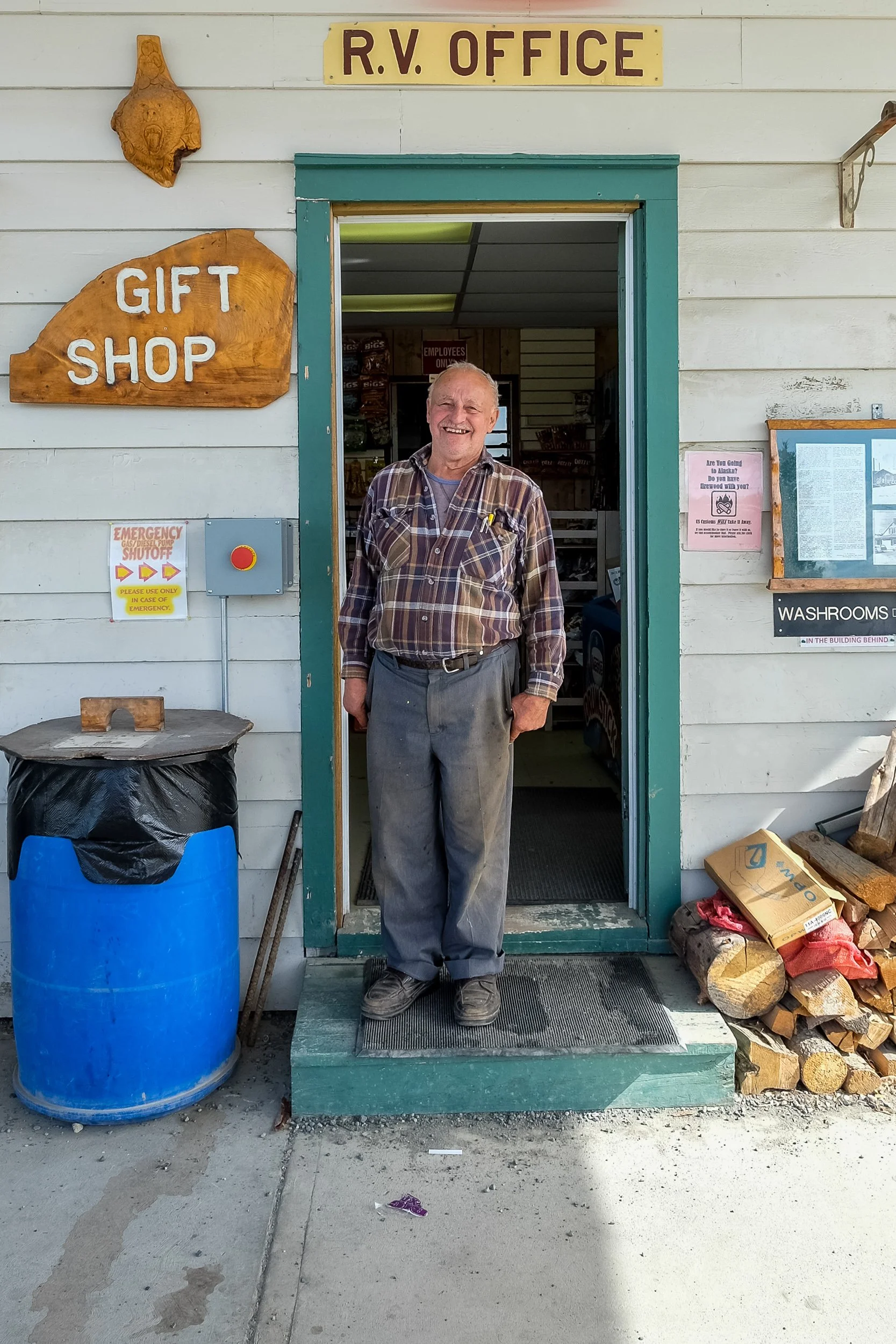 A cheerful elderly man smiling and standing at the entrance of an RV office, wearing a plaid shirt and gray pants. The building has a sign that reads 'R.V. Office' and another that says 'Gift Shop.' There is a large blue barrel, firewood stacked on t