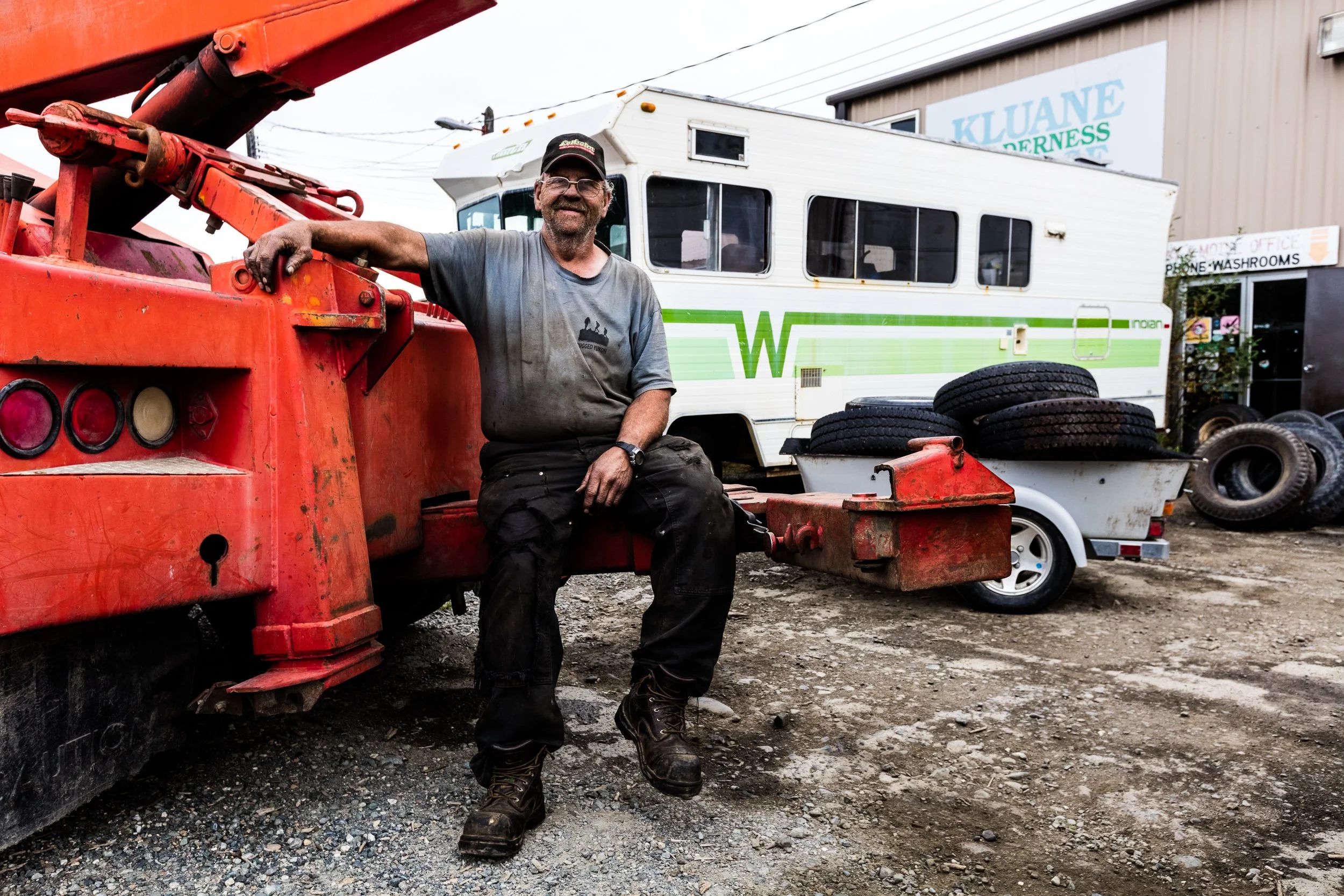 A man wearing a cap, glasses, and a gray T-shirt sitting on a red piece of machinery outdoors, with a white Green Wally camper trailer and tire stacks in the background.