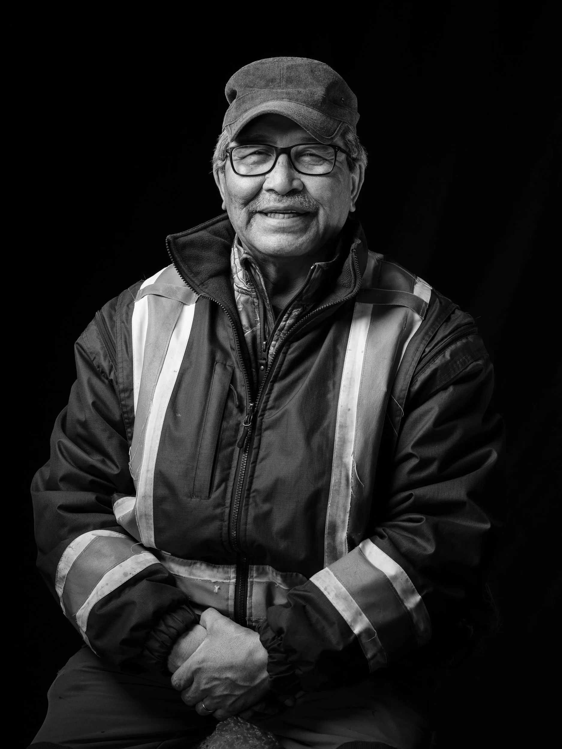 Black and white portrait of an older man smiling, wearing glasses, a cap, a high-visibility jacket, and gloves, against a plain black background.