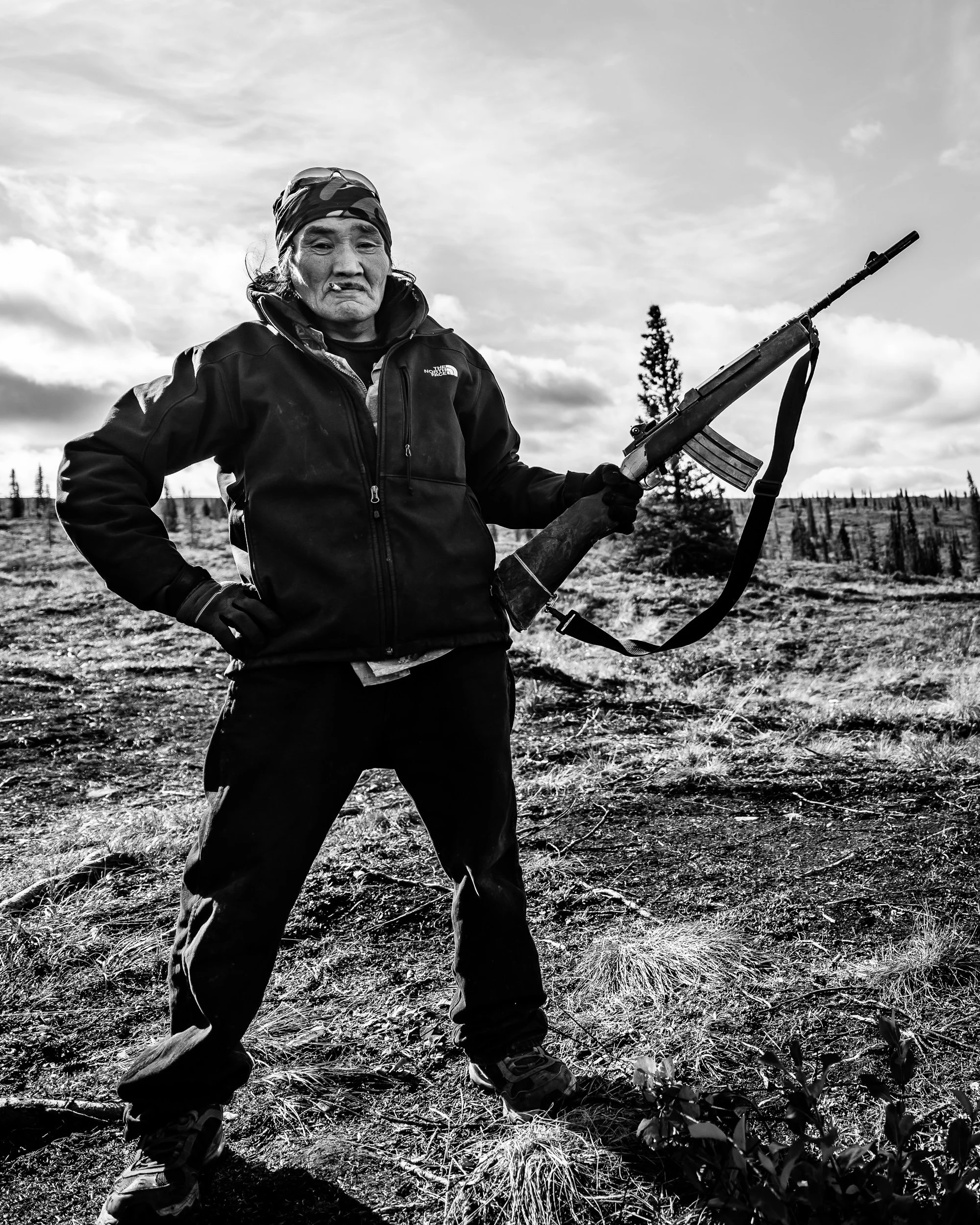 A man dressed in outdoor gear, holding a rifle, standing in a natural landscape with trees and cloudy sky in black and white.