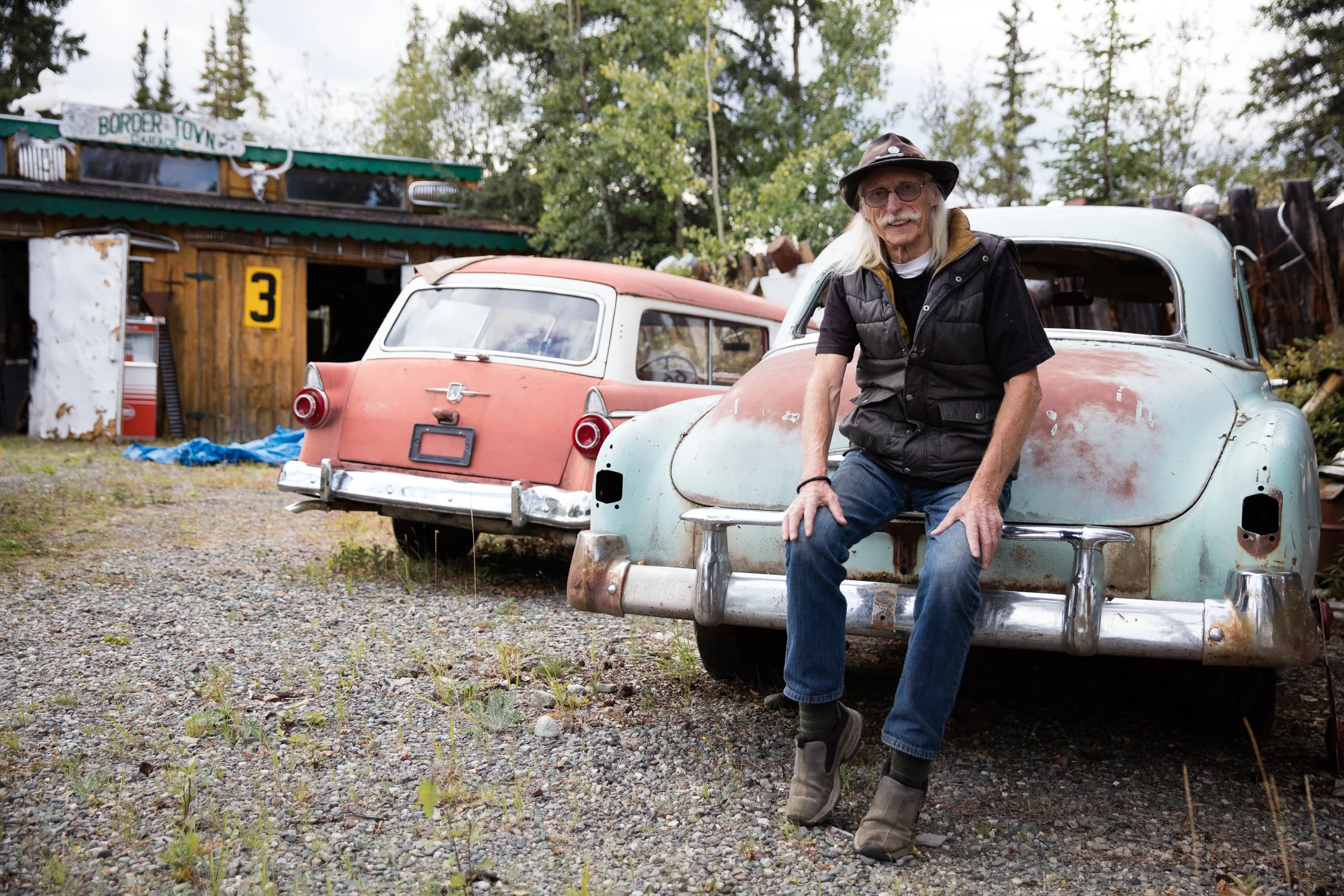 An elderly man with long white hair, glasses, and a mustache, wearing a hat, black vest, and jeans, sitting on the front of a rusty vintage car in a rural outdoor setting.