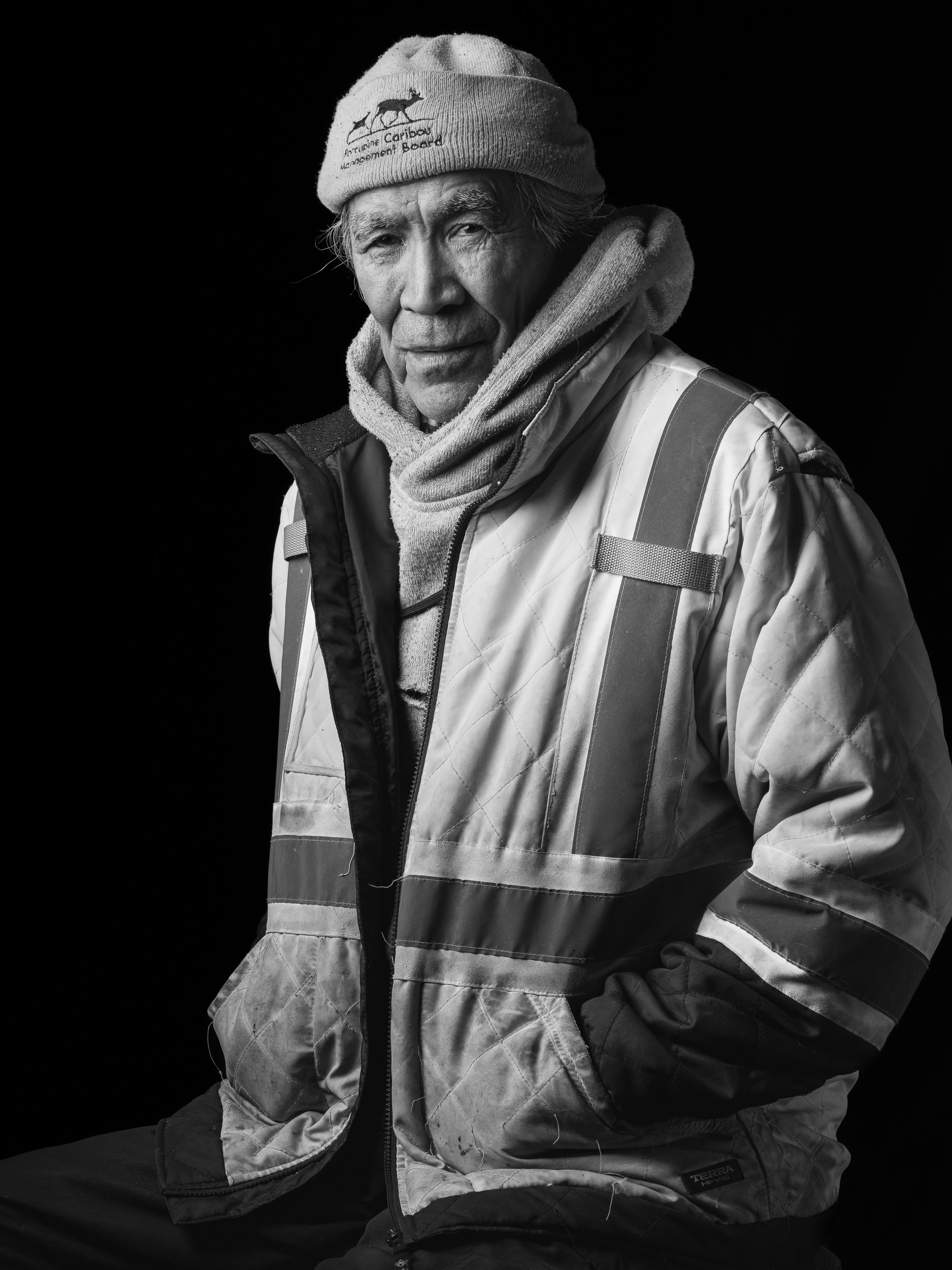 A black and white portrait of an elderly man wearing a knit cap, a hoodie, and a reflective jacket, sitting against a dark background.