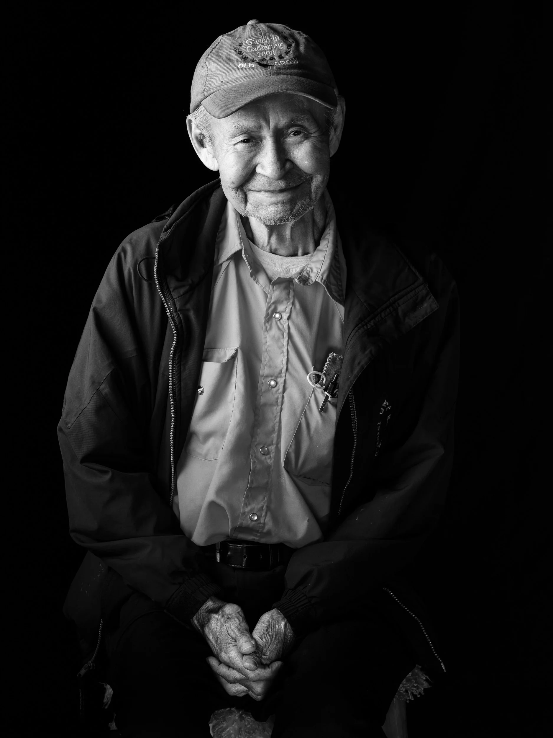 Black and white portrait of an elderly man smiling, wearing a baseball cap, a light-colored collared shirt, and a dark jacket, sitting with hands clasped.