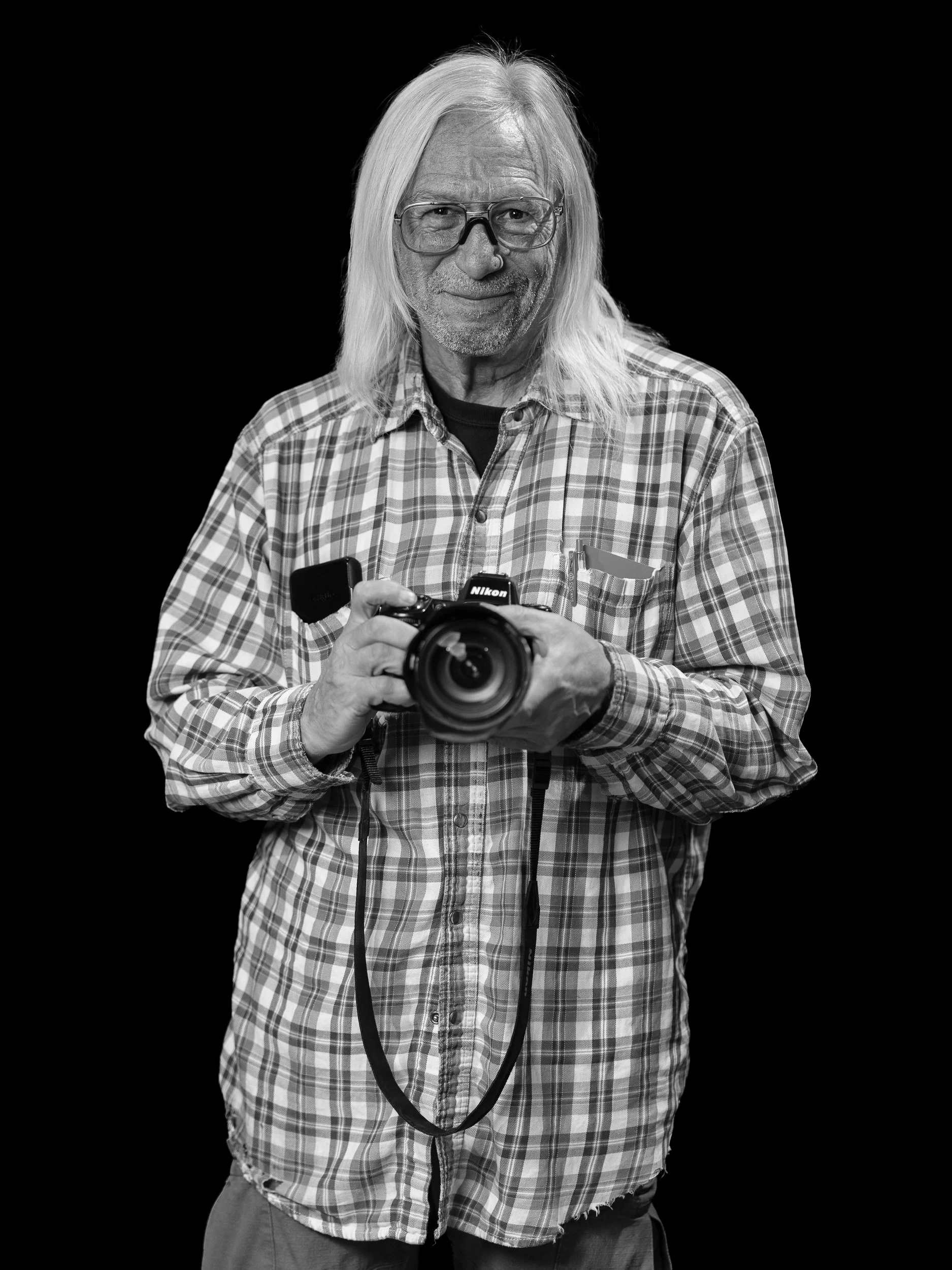Black and white photo of an elderly man with long white hair, glasses, and a slight smile, holding a camera, wearing a checkered shirt.