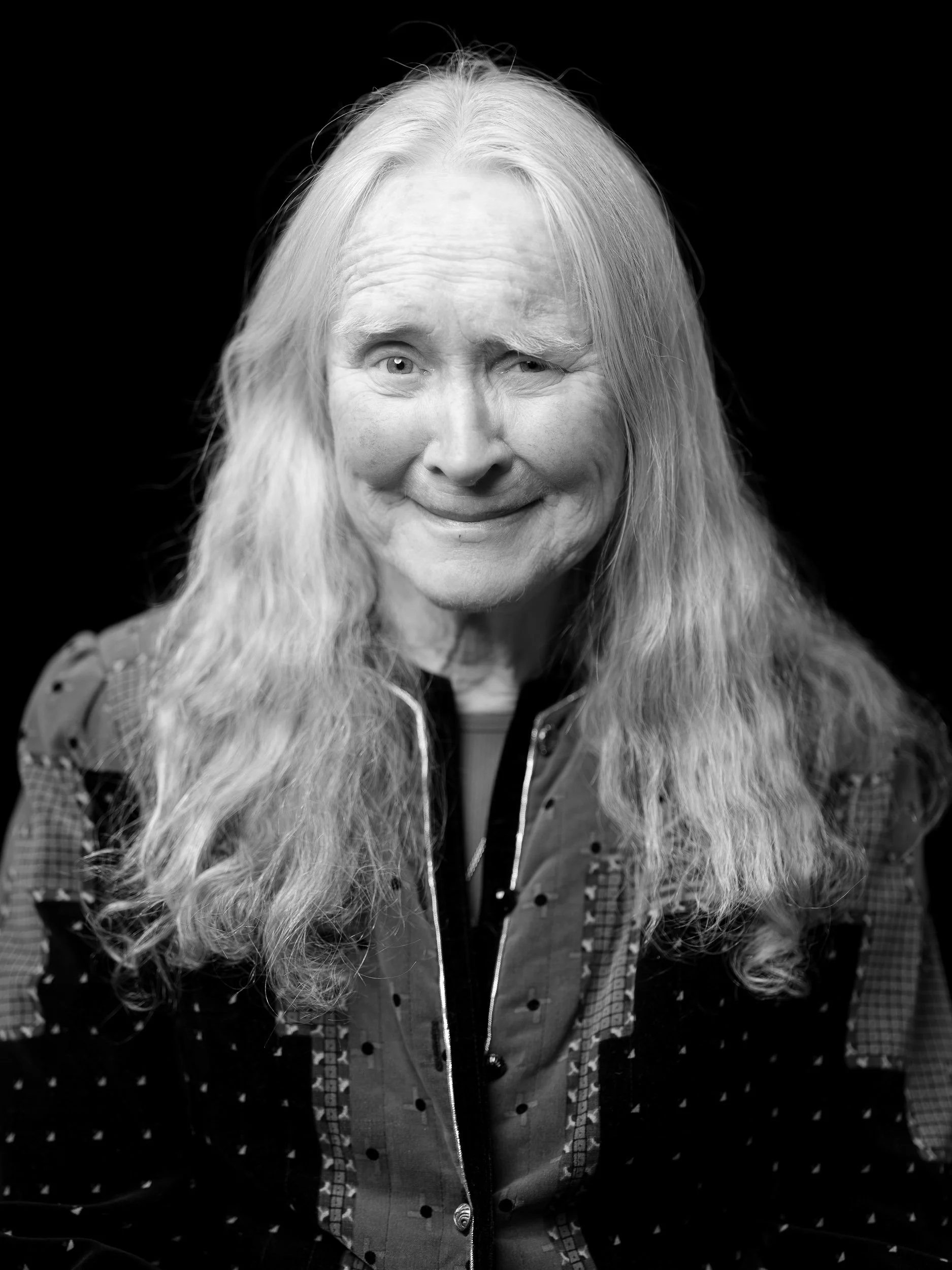 Black and white portrait of an elderly woman with long, wavy hair, smiling and winking. She is wearing a patterned jacket with a zipper.
