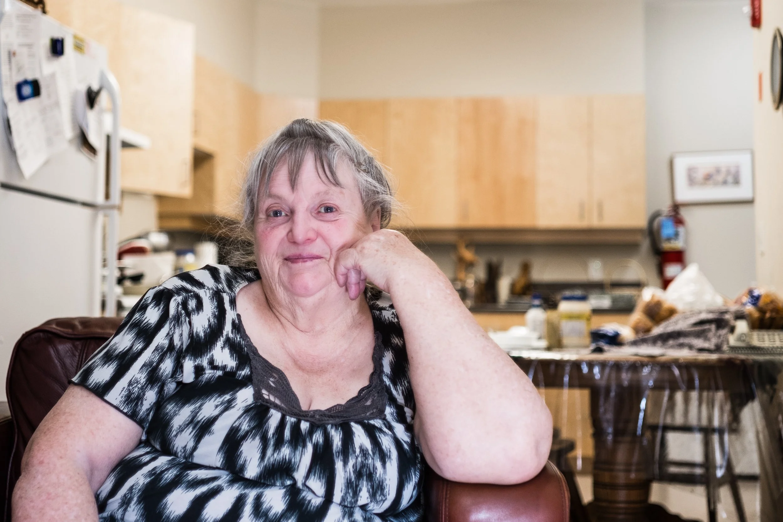An elderly woman with gray hair and a black and white patterned top sitting in a kitchen, resting her face on her hand and smiling gently.