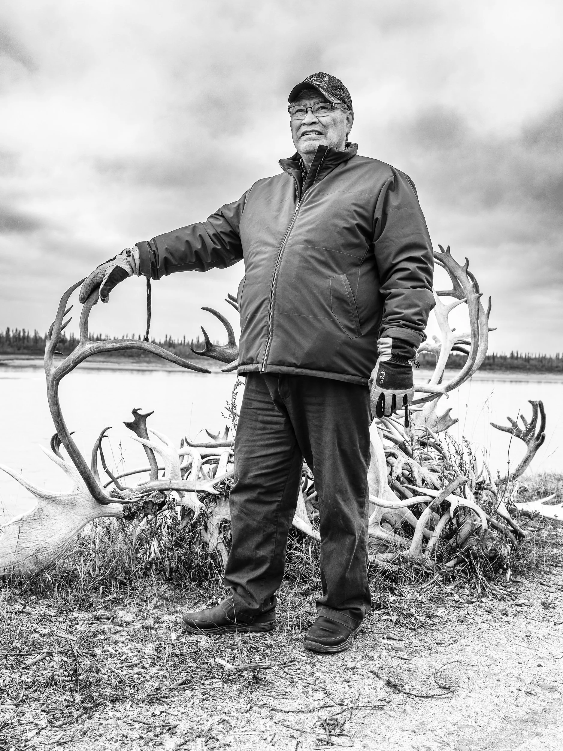Black and white photo of an older man holding large antlers in a natural outdoor setting with a cloudy sky, wearing glasses, a cap, a jacket, and gloves.