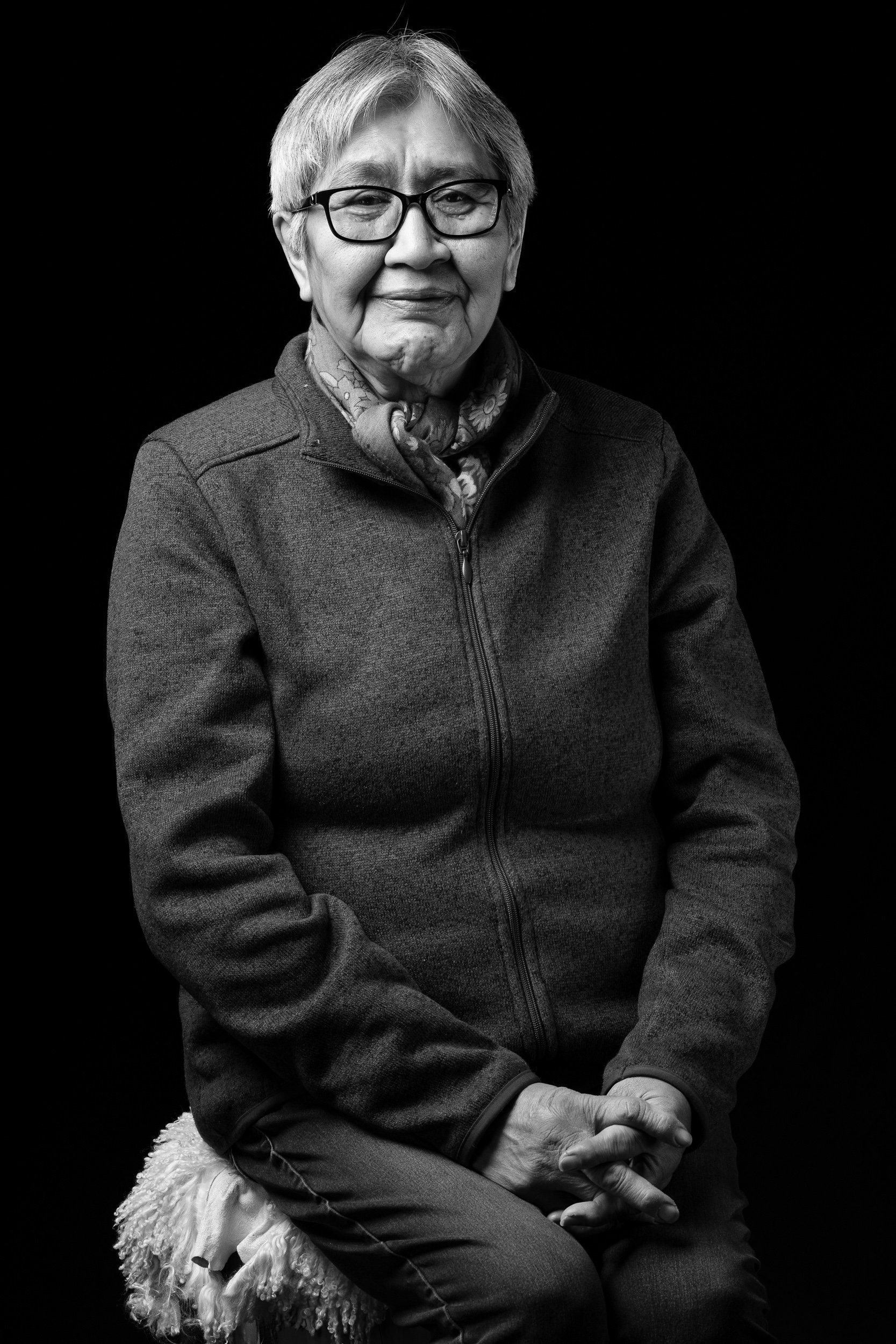 Black and white portrait of an elderly woman with short light-colored hair and glasses, sitting with hands clasped, smiling slightly, wearing a zip-up jacket and scarf against a black background.