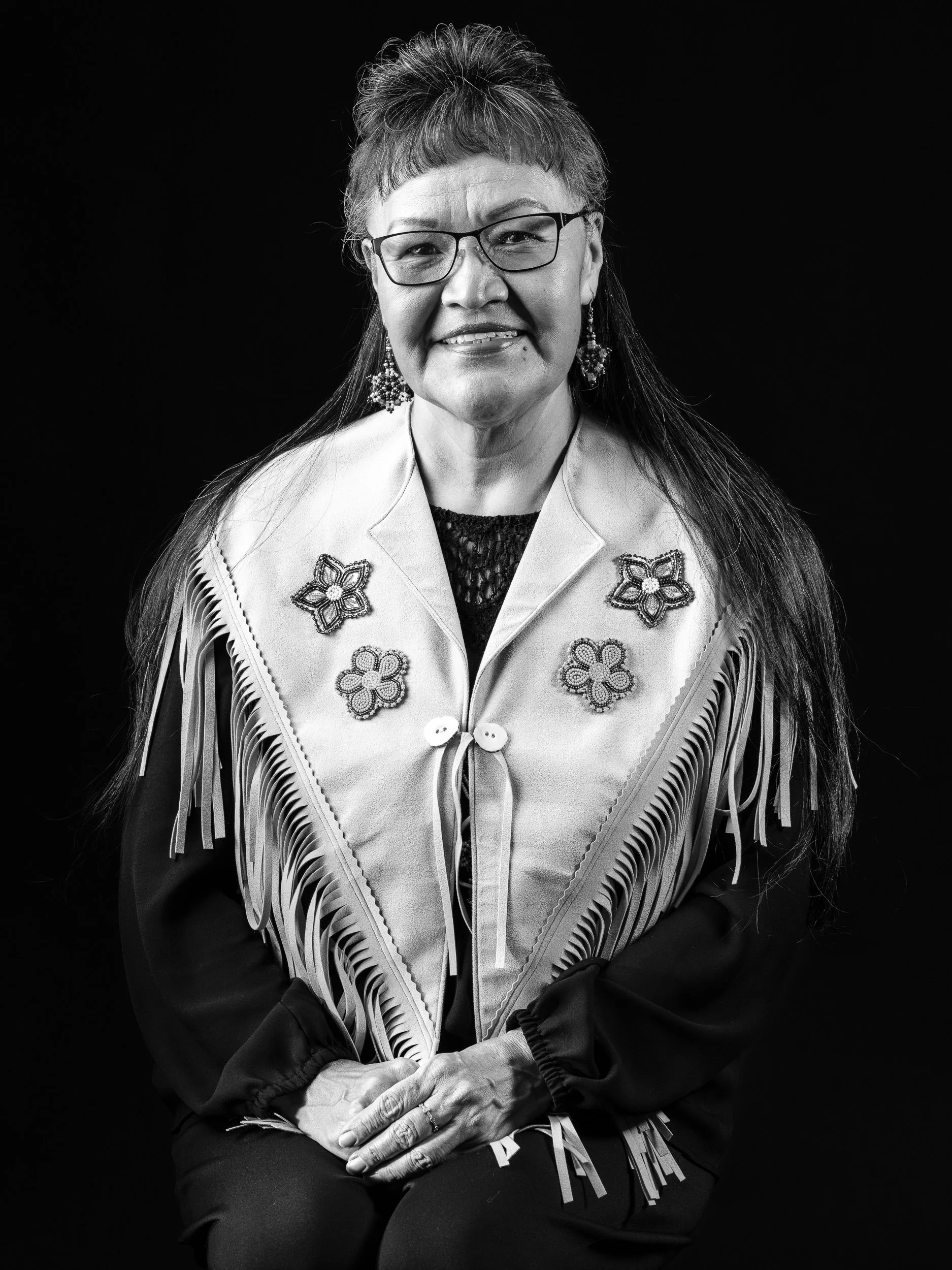 Black and white portrait of a woman wearing glasses, earrings, a decorated shawl with floral patterns, and fringes, sitting against a black background.