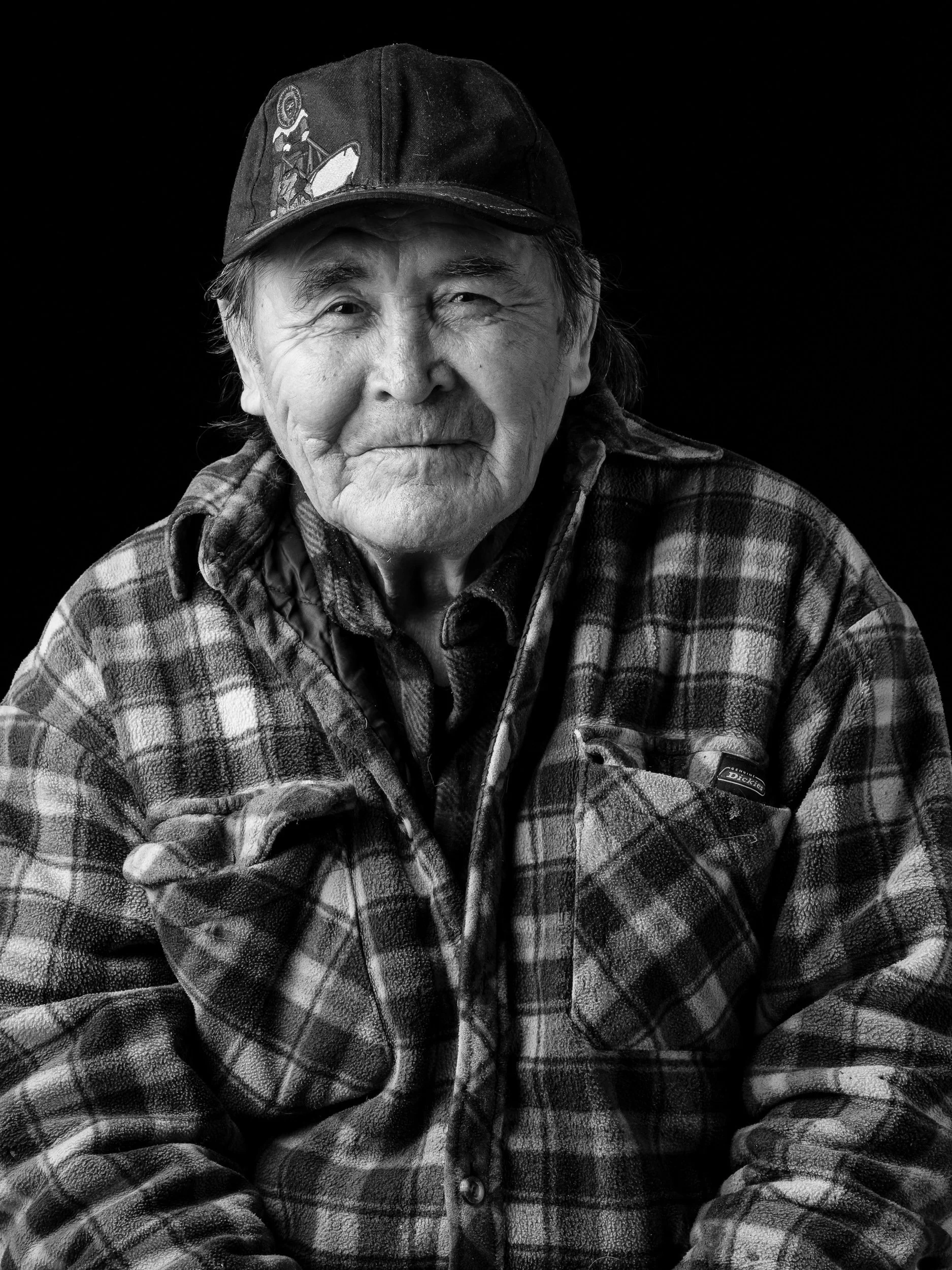 Black and white portrait of an elderly man wearing a baseball cap and a plaid jacket, smiling softly against a dark background.