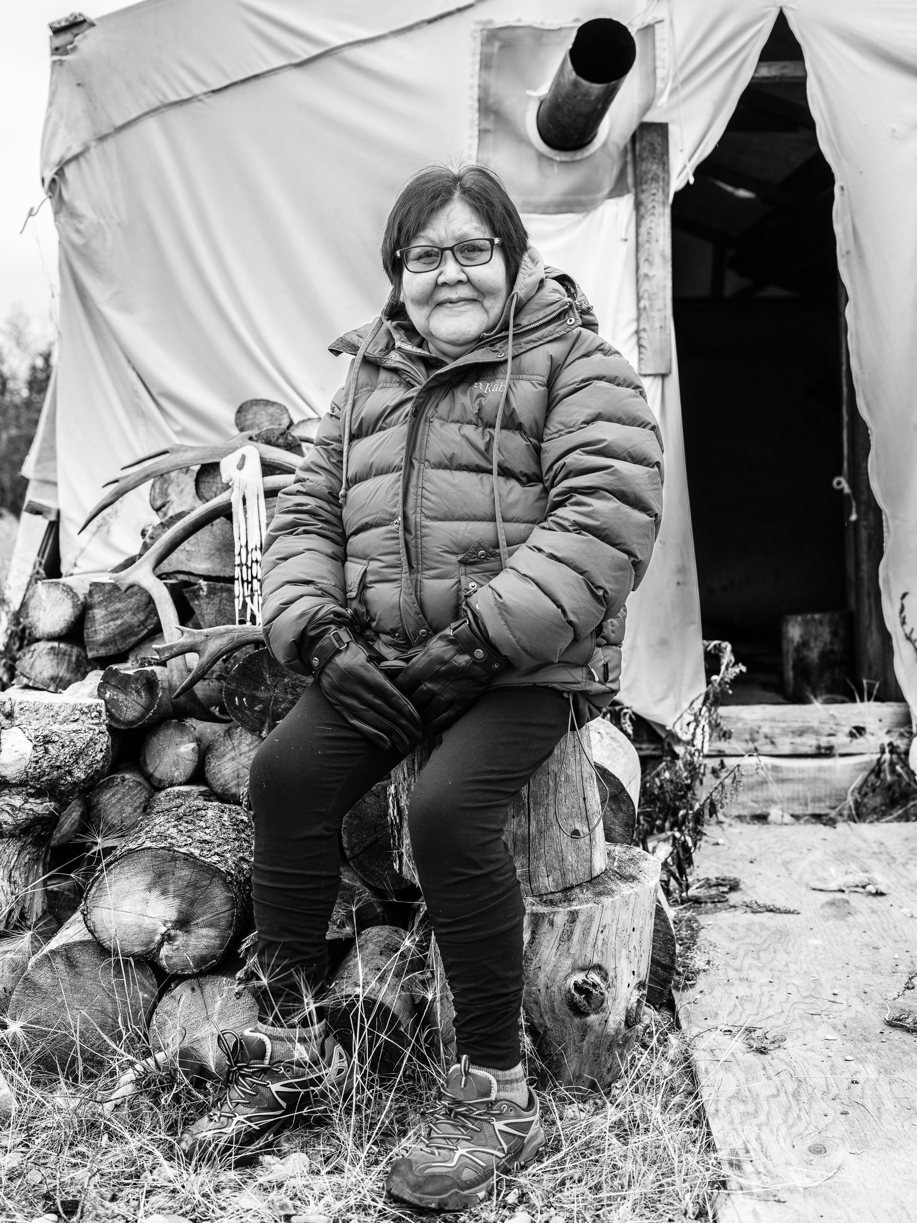 A woman sitting outdoors on a log in front of a pile of firewood and a small tarp shelter, wearing glasses, a padded jacket, gloves, and hiking boots.
