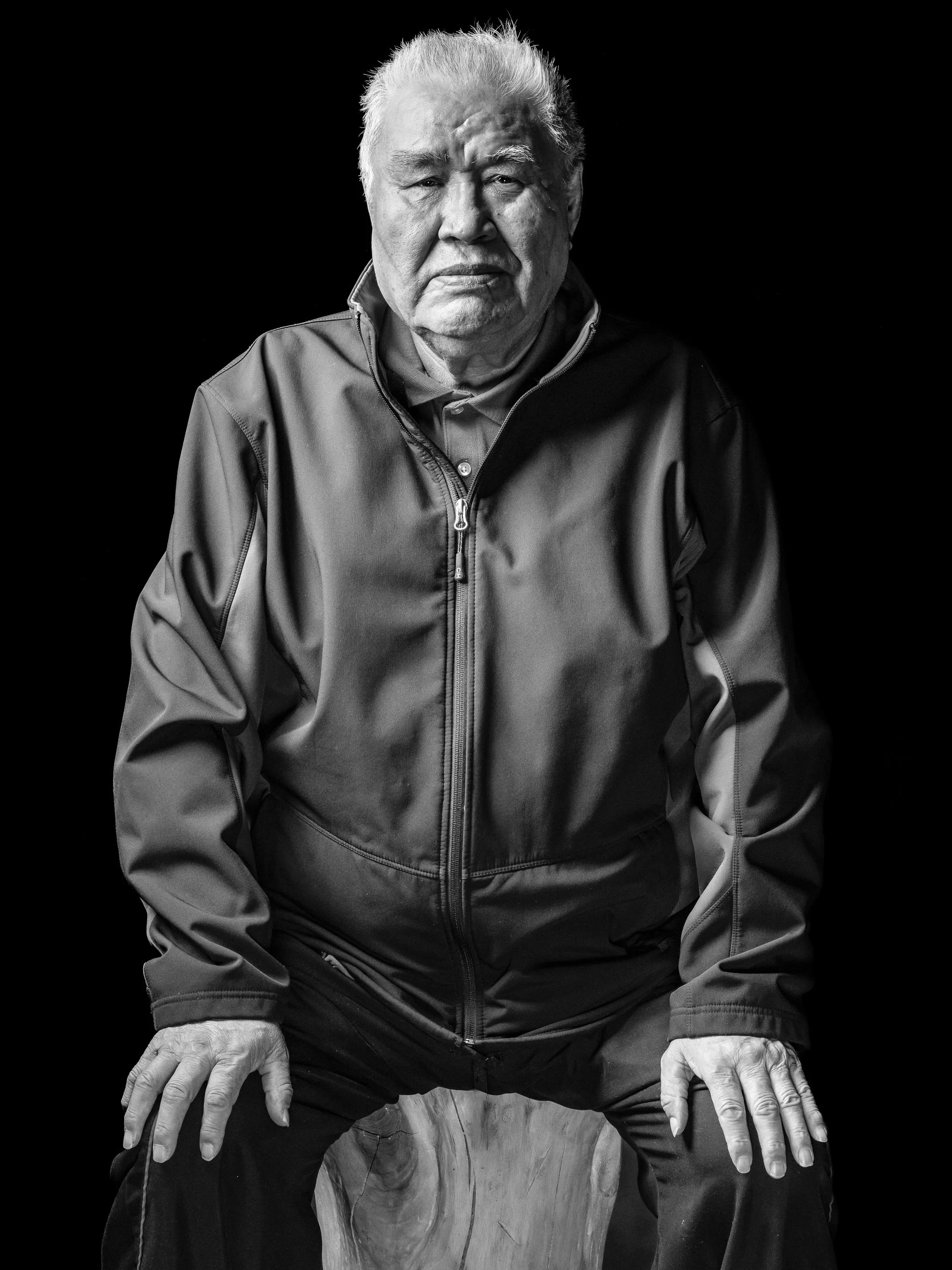 Black and white portrait of an elderly man with white hair, wearing a zip-up jacket, sitting on a wooden stool, with his hands resting on his knees, against a dark background.