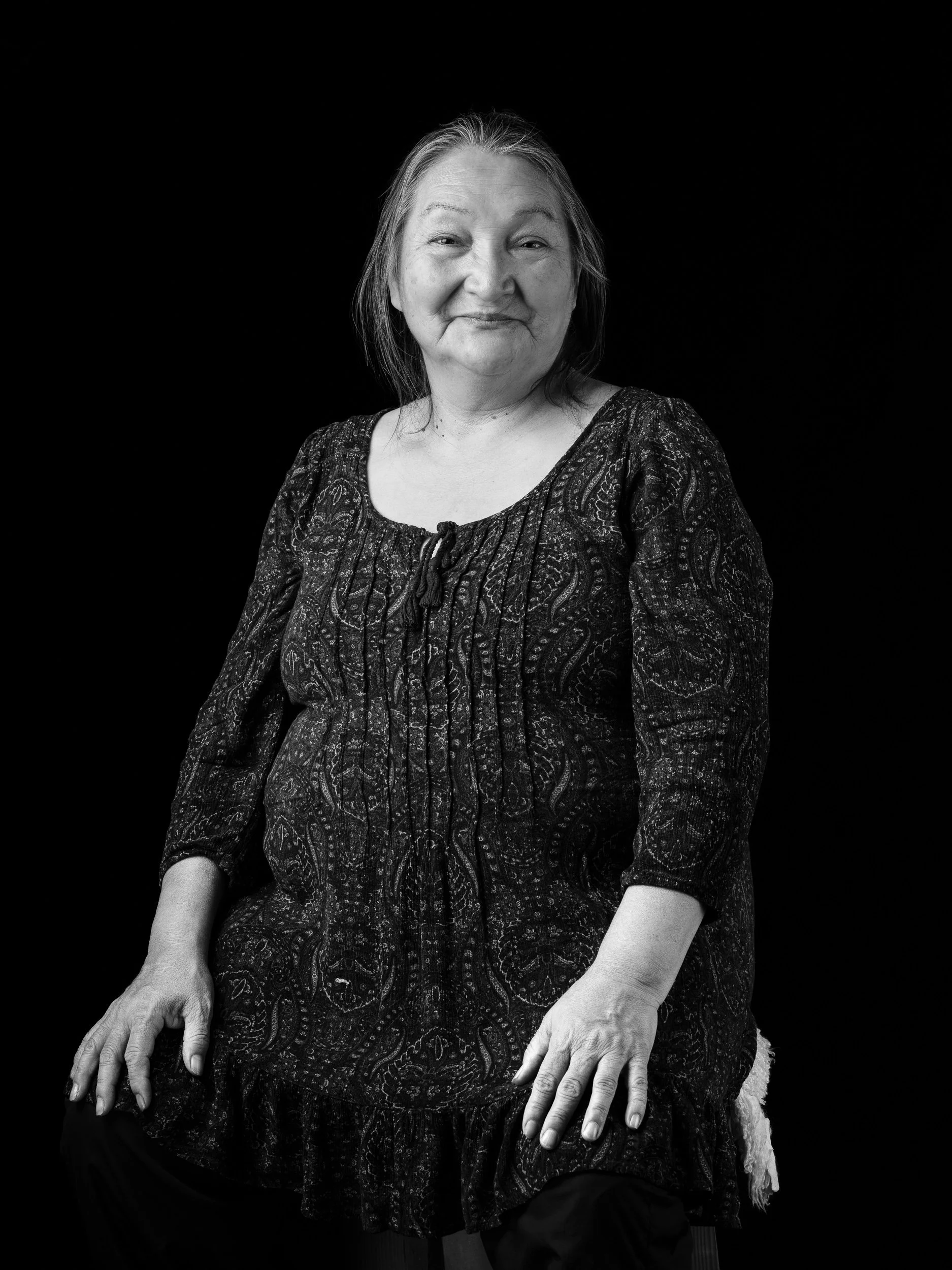 Black and white photo of an elderly woman smiling, sitting against a plain black background, wearing a patterned dress.