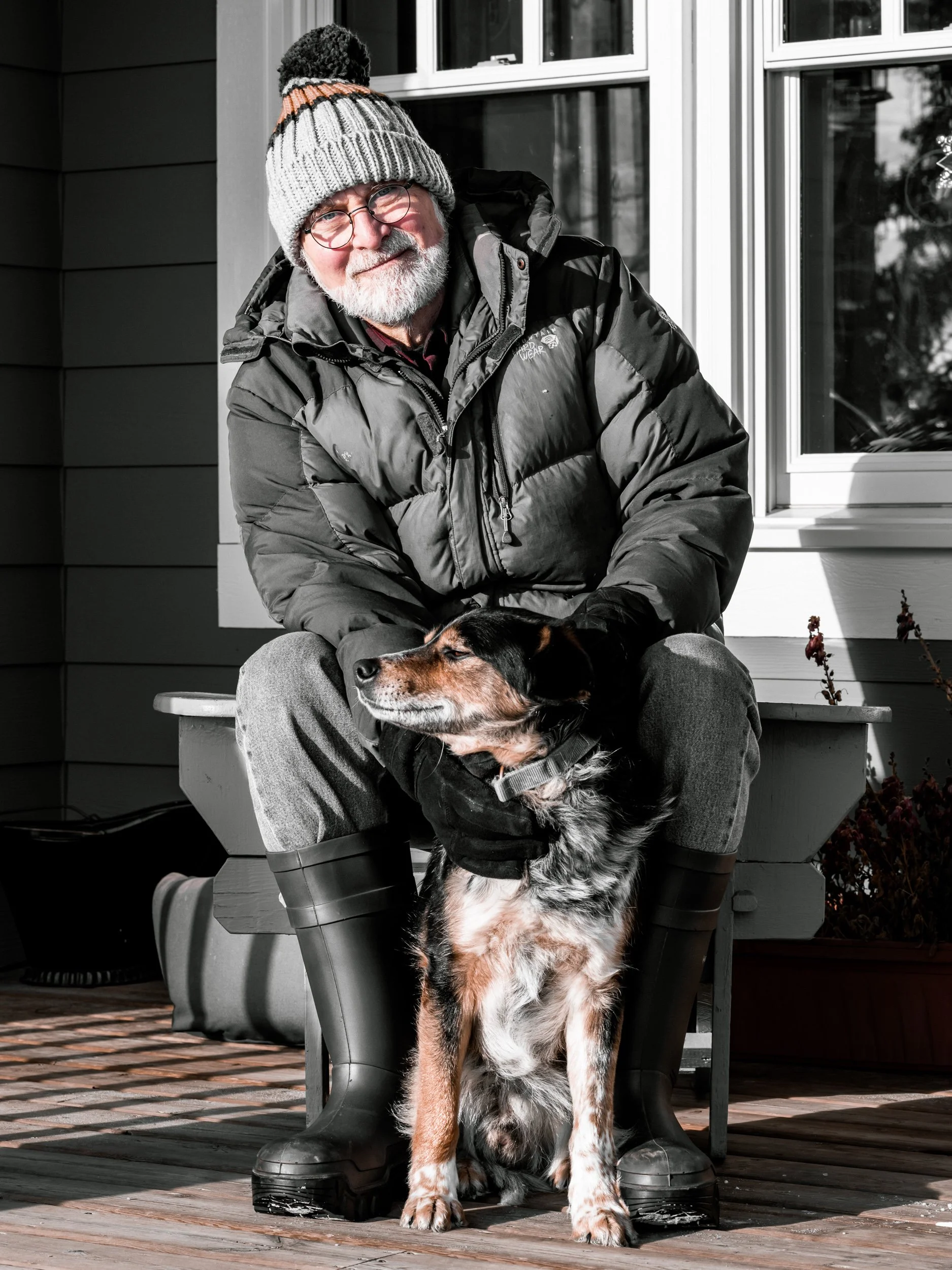 An elderly man with glasses, a gray beard, and a knit winter hat sitting on a bench outdoors with a large dog. The man is wearing a puffy jacket and gray pants, and is smiling while looking at the camera.