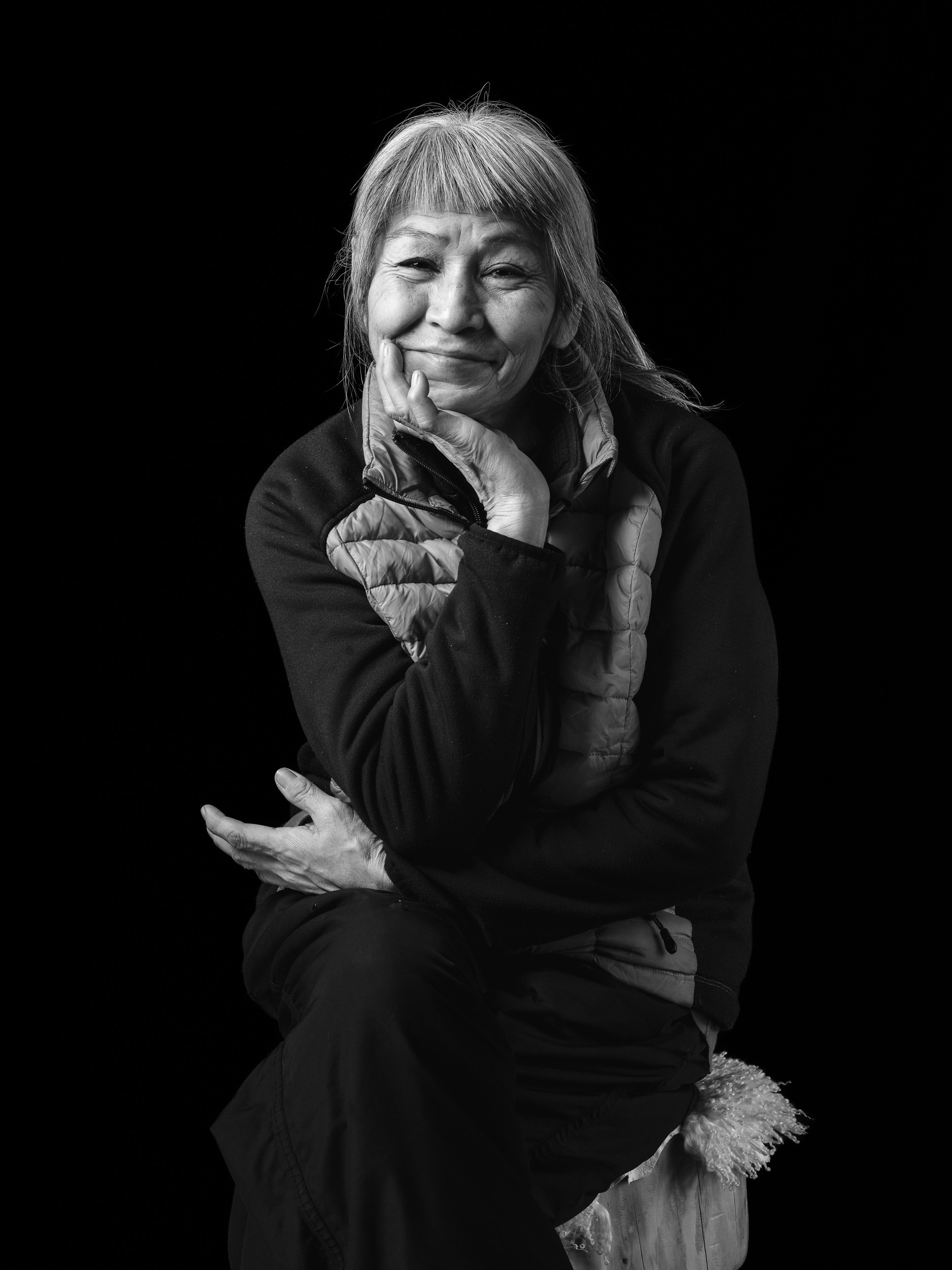 Black and white portrait of an elderly woman smiling, with her hand resting on her cheek, wearing a jacket with a high collar, and sitting on a wooden surface with a furry cushion.