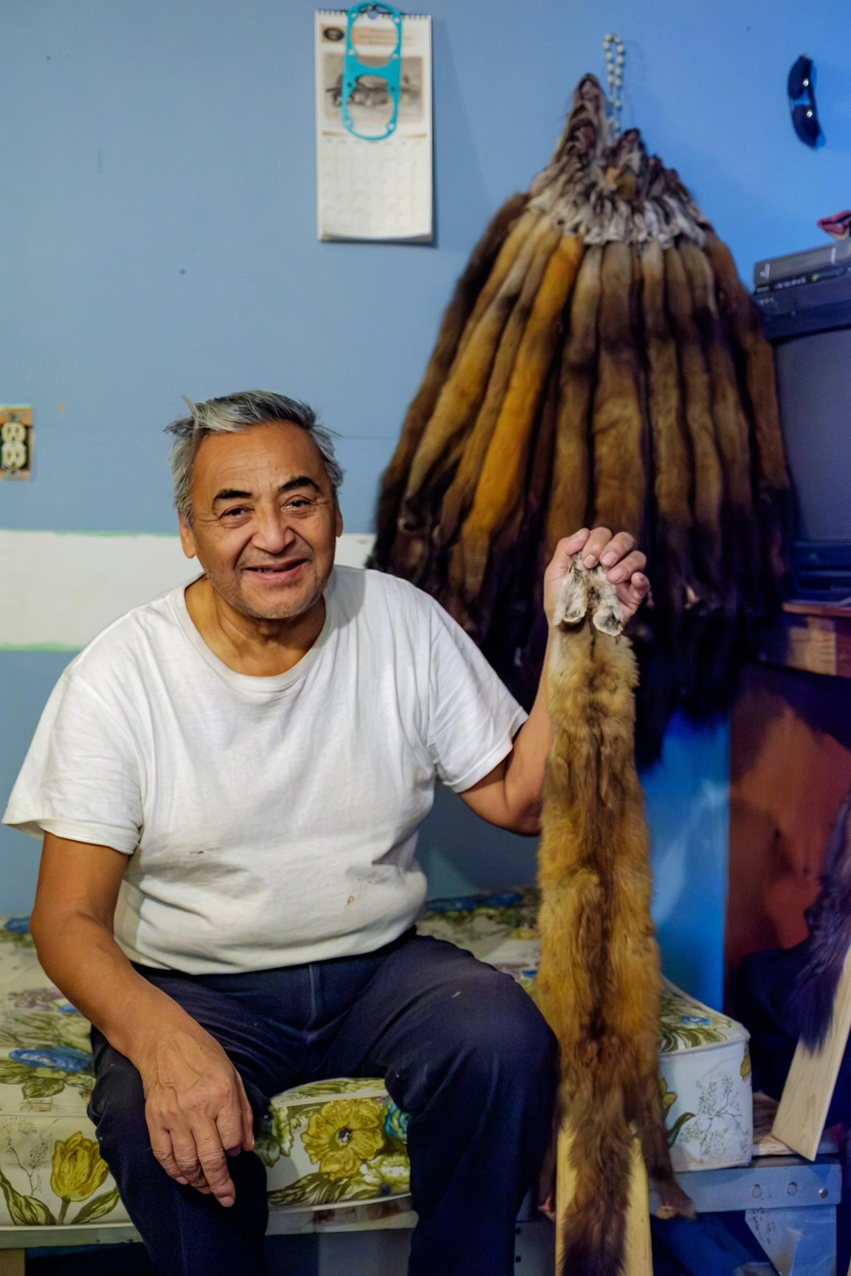 An elderly man with gray hair smiling, sitting on a floral patterned bench, holding a large feline paw attached to a taxidermy fox mount that is facing upward. The background includes a blue wall with a calendar and some objects on a shelf.