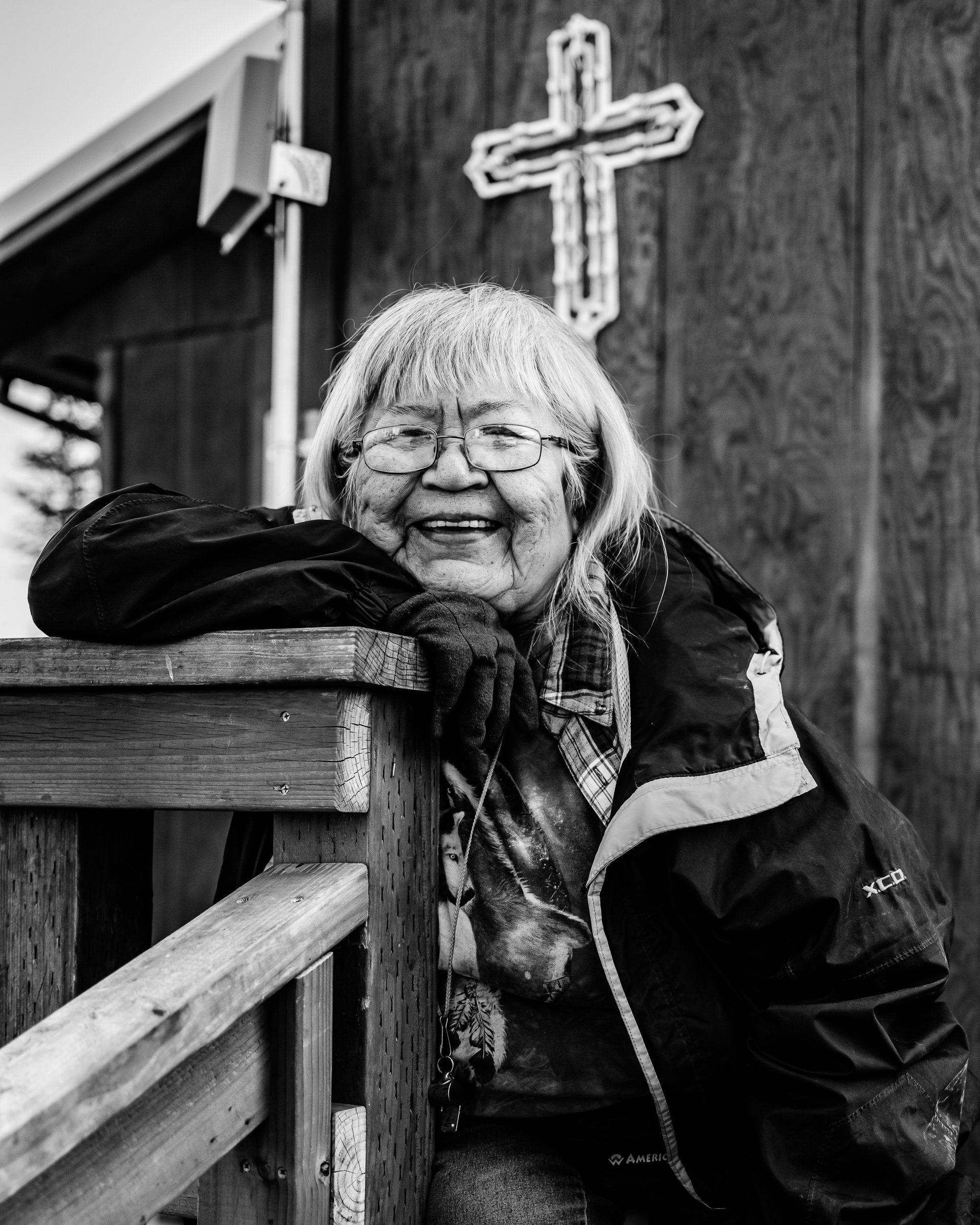 An elderly woman with glasses, light-colored hair, and wearing a jacket leaning on a wooden railing outside with a cross sign on the dark wooden wall behind her.